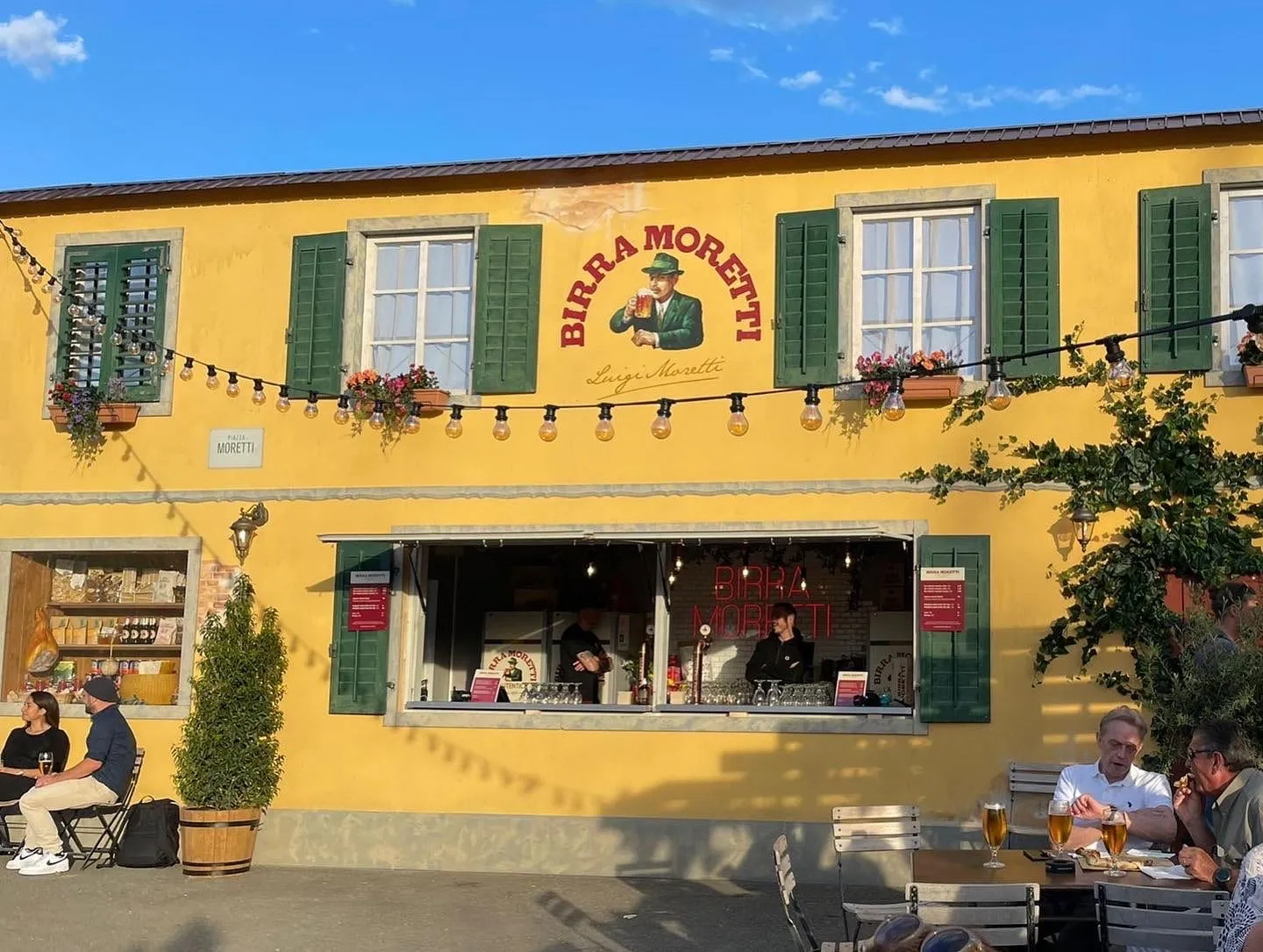 Birra Moretti bar built as a yellow house facade with green shutters, a string of lights and guests at tables