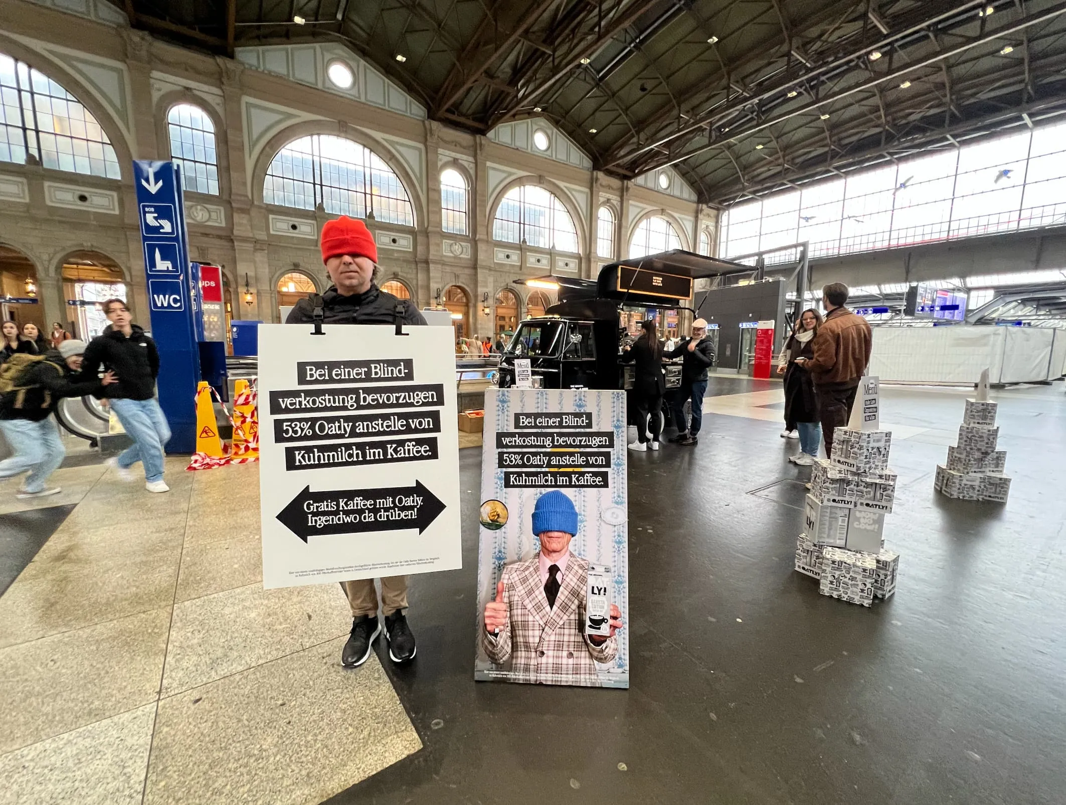 Promoter with a red hat over his eyes and an Oatly sandwich board in the station hall, beside a coffee Piaggio and Oatly cartons