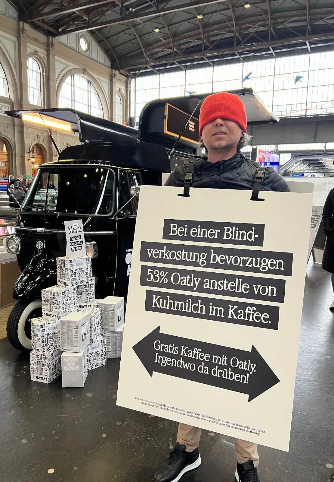 Sandwichman mit roter Mütze über den Augen und Oatly-Schild vor dem schwarzen Kaffee-Piaggio mit Oatly-Kartons
