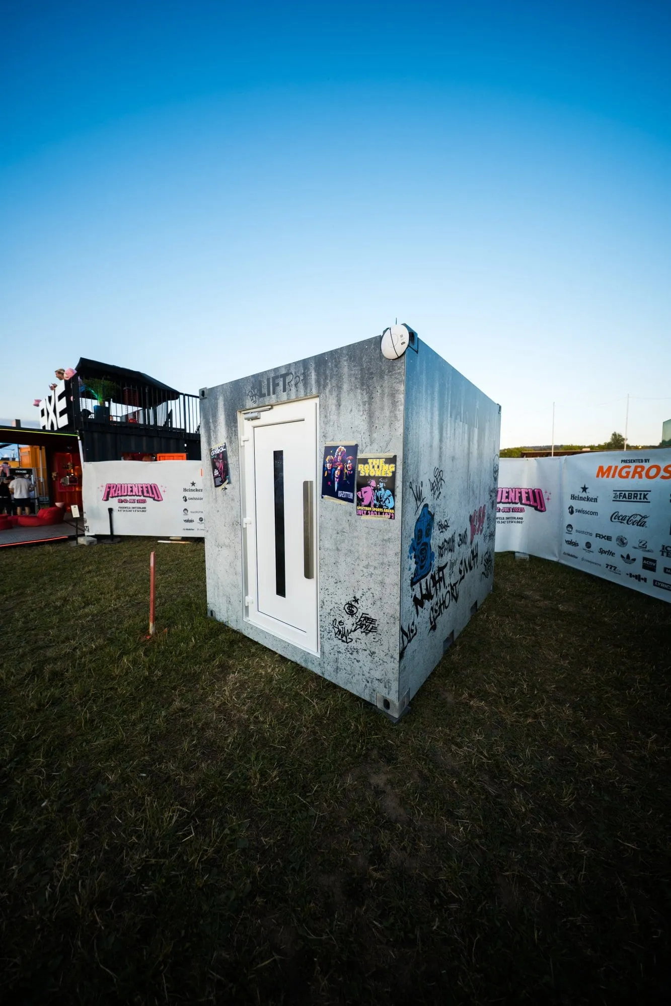 Betonlook-Container mit weisser Lifttür, Konzertpostern und Graffiti auf der Festivalwiese, dahinter Sponsorenbanner