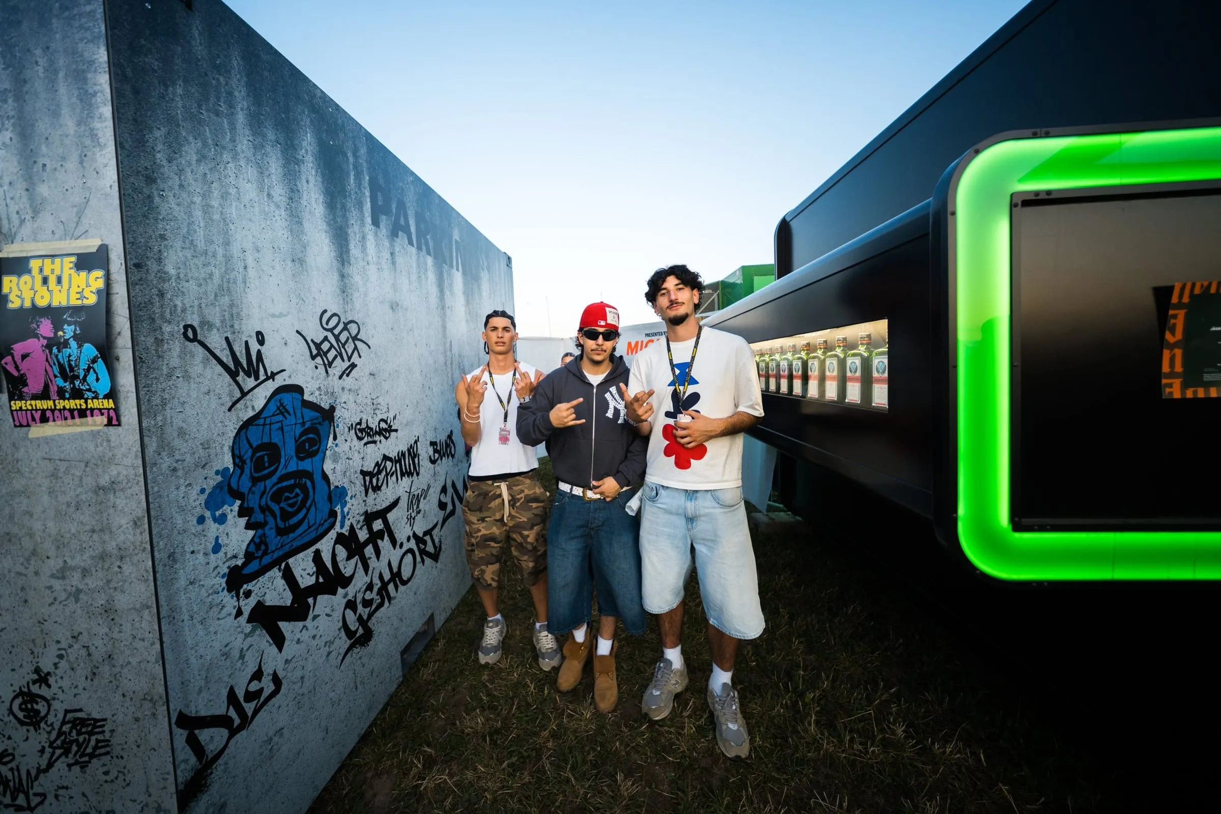 Drei junge Männer posieren zwischen der Graffiti-Wand des Containers und einer schwarzen Bar mit grünem Leuchtrahmen und Jägermeister-Flaschen