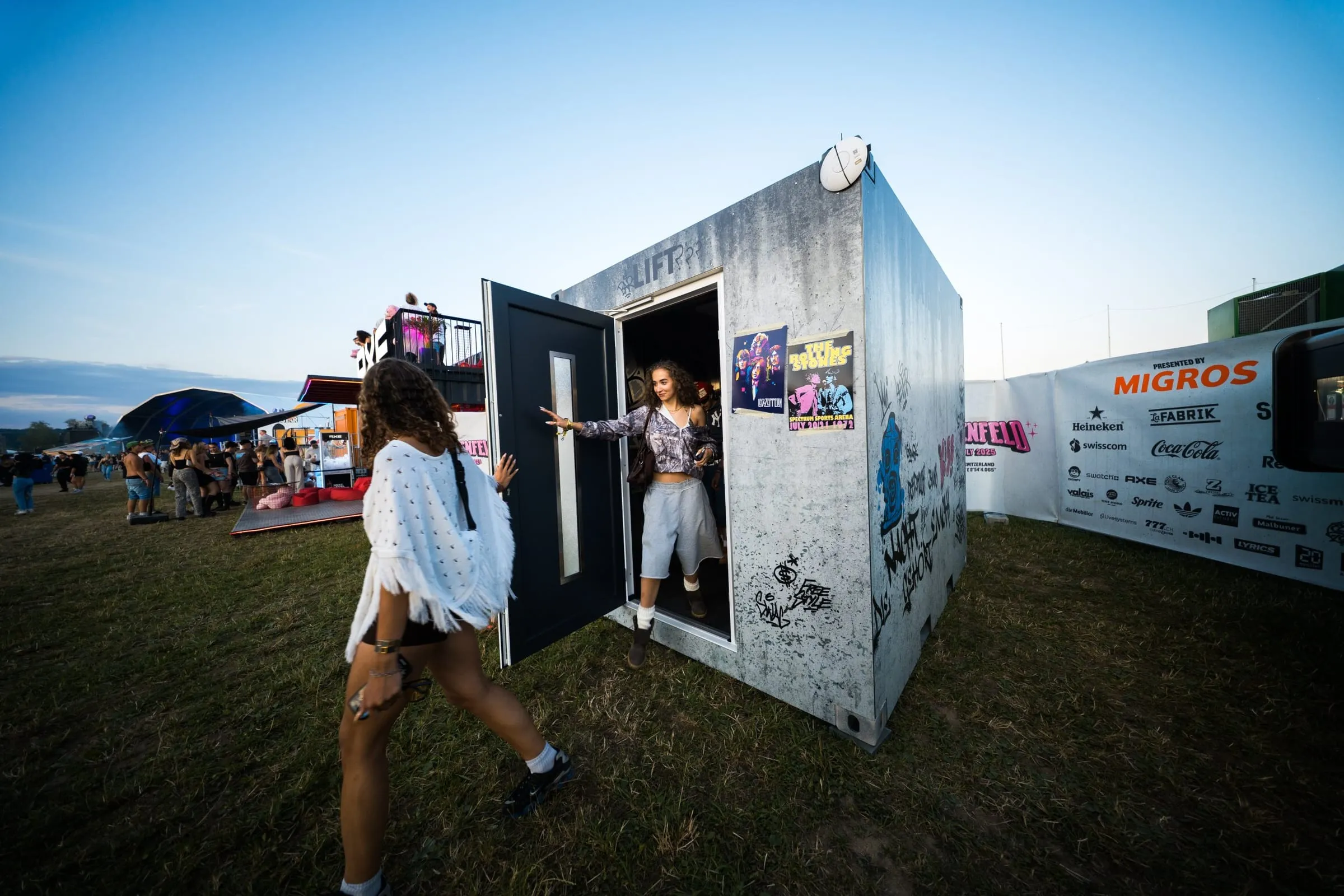 Concrete-look container with graffiti and «Lift» lettering, two visitors at the open door, with the festival site at dusk