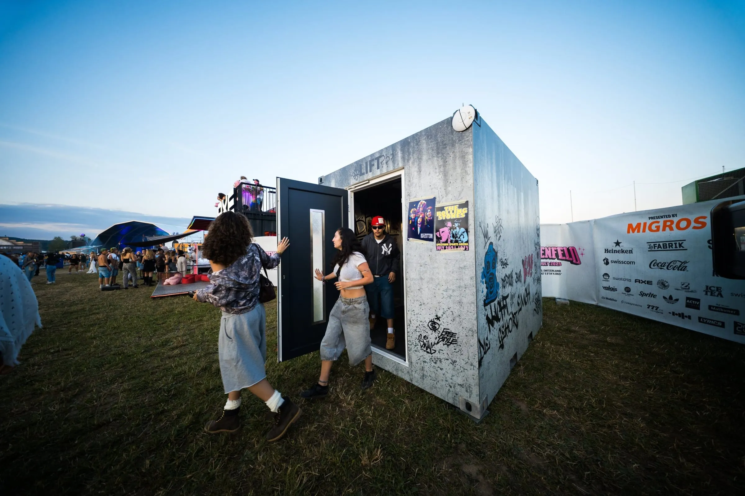 Zwei Besucherinnen öffnen die dunkle Tür des Lift-Containers, ein Mann mit roter Cap tritt heraus