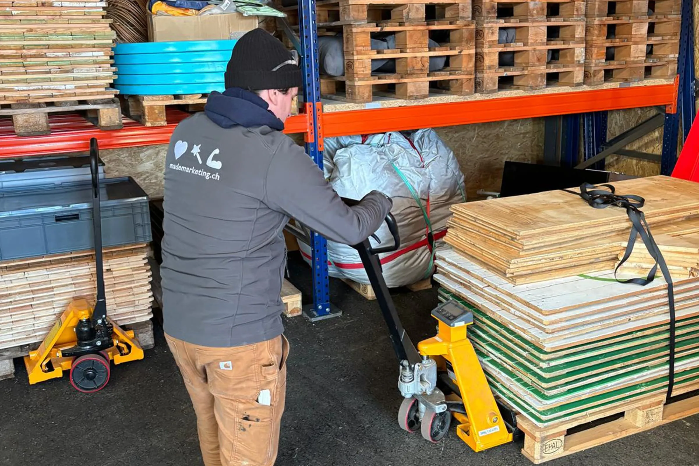 Man in a grey made Marketing jacket pushing a pallet truck under a stack of wooden boards in the store