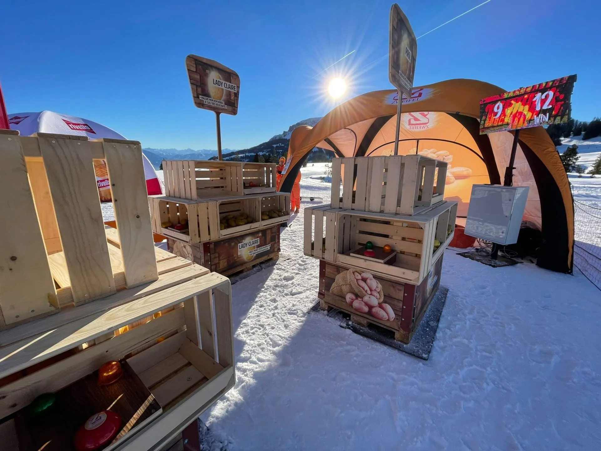 Wooden crates with potatoes, buzzers and variety labels in the snow, in front of the orange Zweifel tent and scoreboard