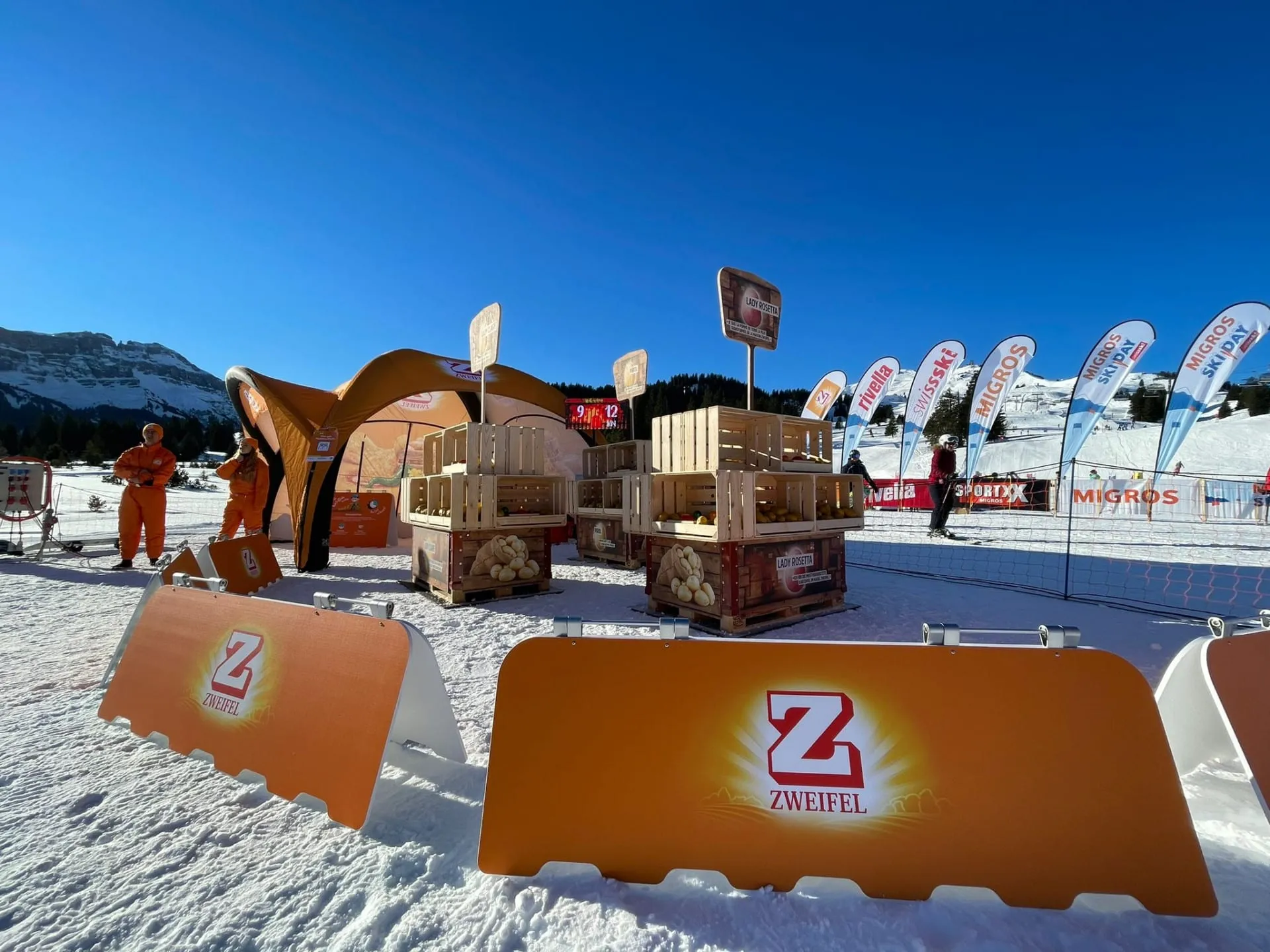 Orange Zweifel-Banden und Spielfeld mit Holzkisten im Schnee, Zelt, Promotoren in Orange und Migros-Ski-Day-Fahnen vor Bergkulisse