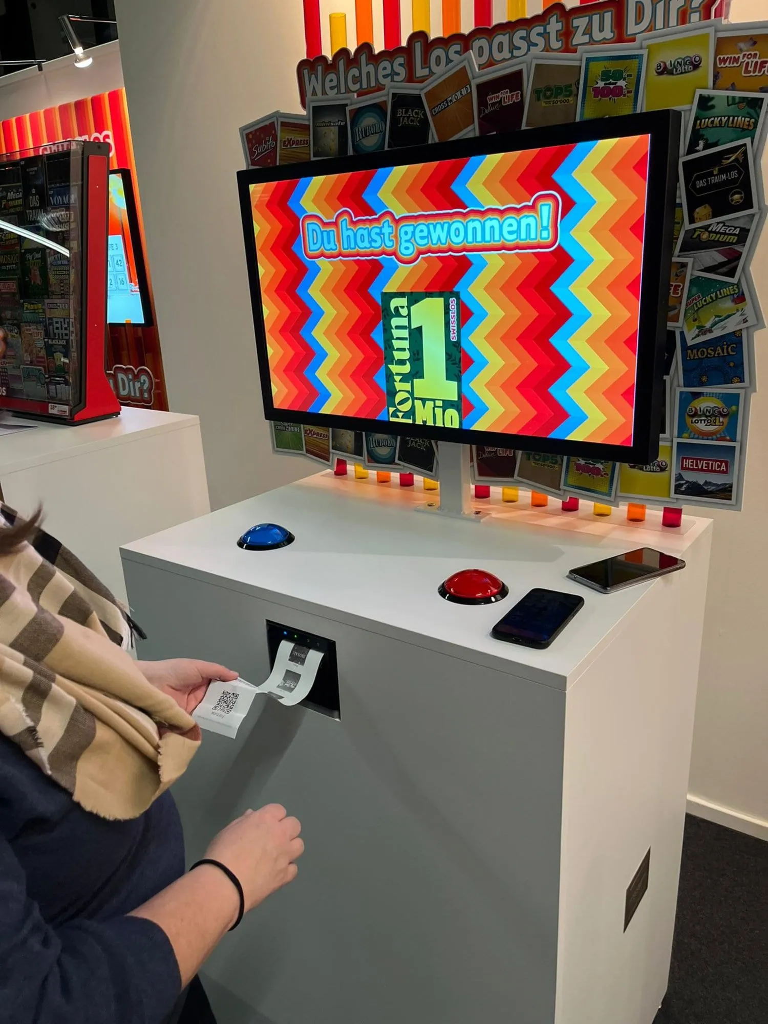 Game-Station mit Bildschirm «Du hast gewonnen!», blauem und rotem Buzzer, Frau zieht Bon aus dem Drucker