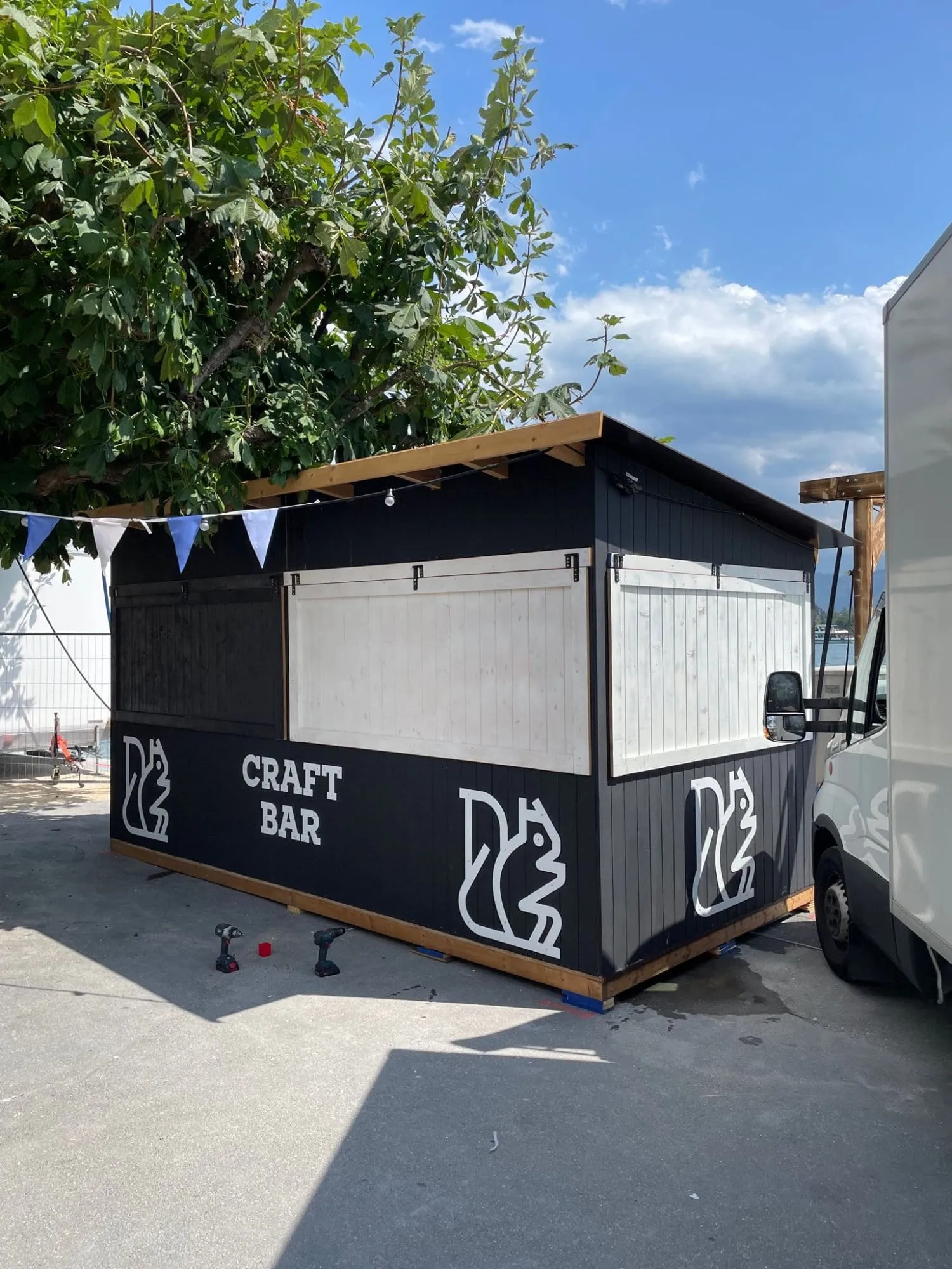 Schwarze Craft Bar mit weissen Eichhörnchen-Logos, hellen Holzklappen und Wimpelkette neben einem Lieferwagen