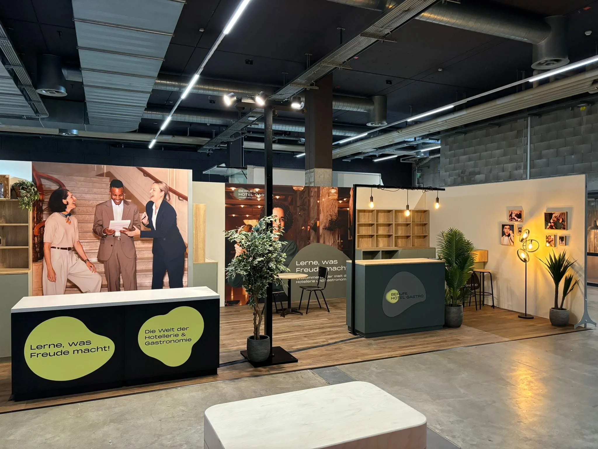 Stand with a «Lerne, was Freude macht!» counter, a photo wall showing three people on stairs, shelves and plants