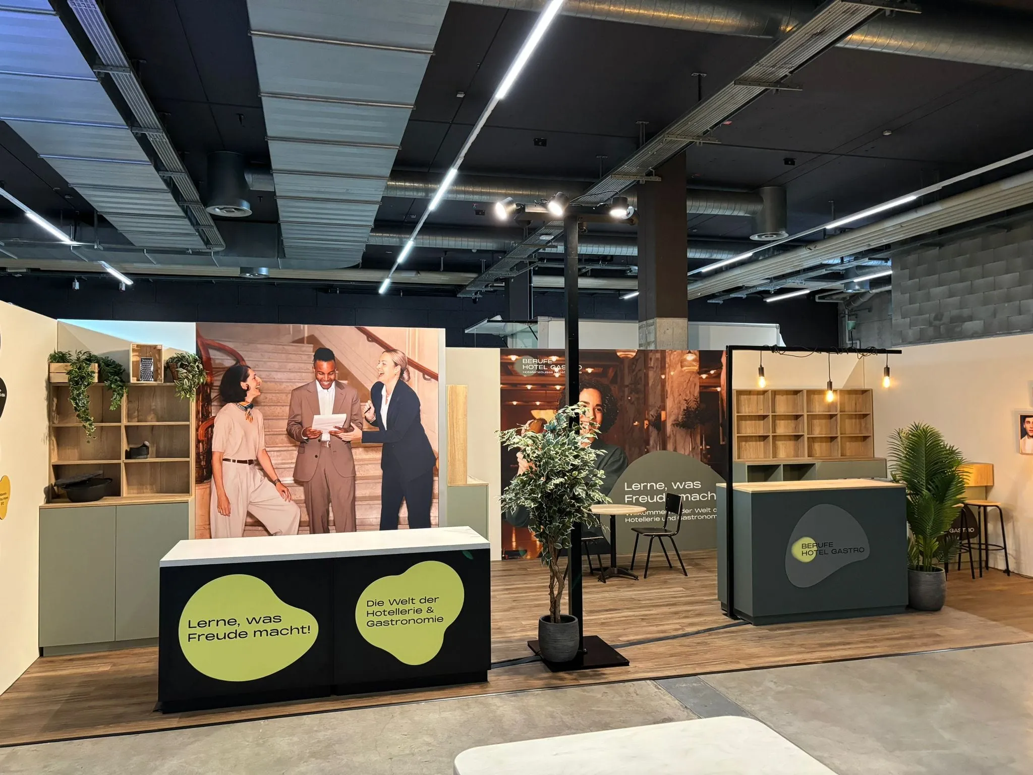 Messestand mit Theke «Lerne, was Freude macht!», grosser Fotowand, Holzregal, Hängelampen und Pflanzen in der Messehalle