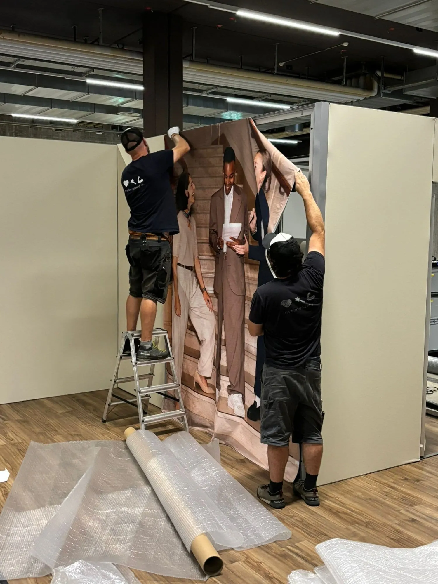 Zwei Männer spannen den Stoffdruck mit dem Treppenfoto auf den Wandrahmen, einer auf der Leiter