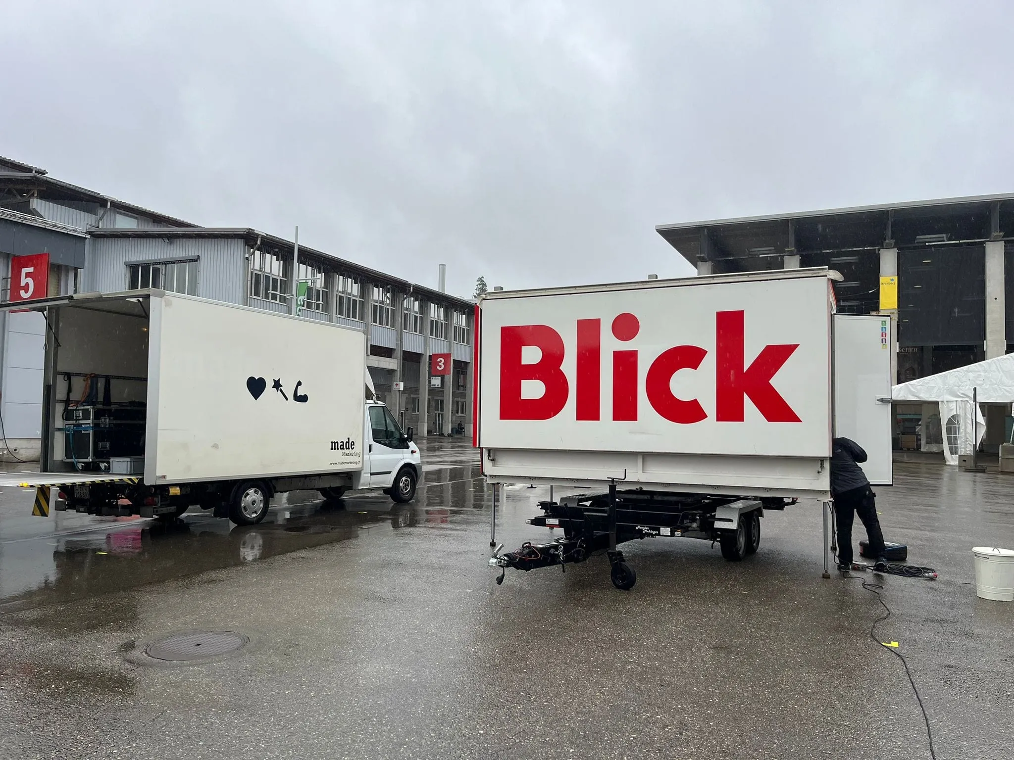 Anhänger mit grossem rotem Blick-Logo neben dem Lieferwagen von made Marketing auf dem nassen Messegelände