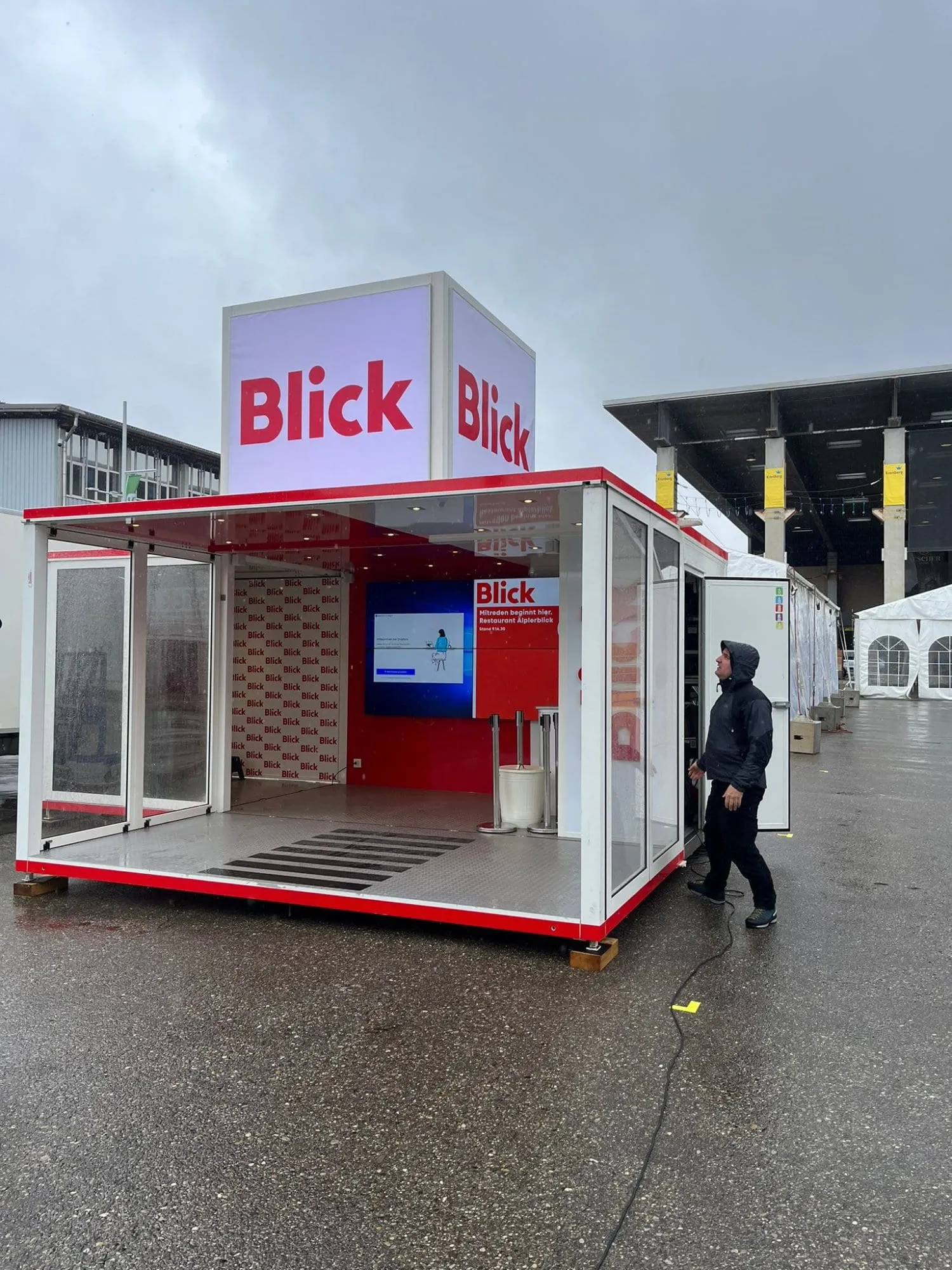 Offene Blick-Box mit Leuchtwürfel auf dem Dach, Blick-Tapete und Bildschirm im Innern, davor ein Mann im Regen