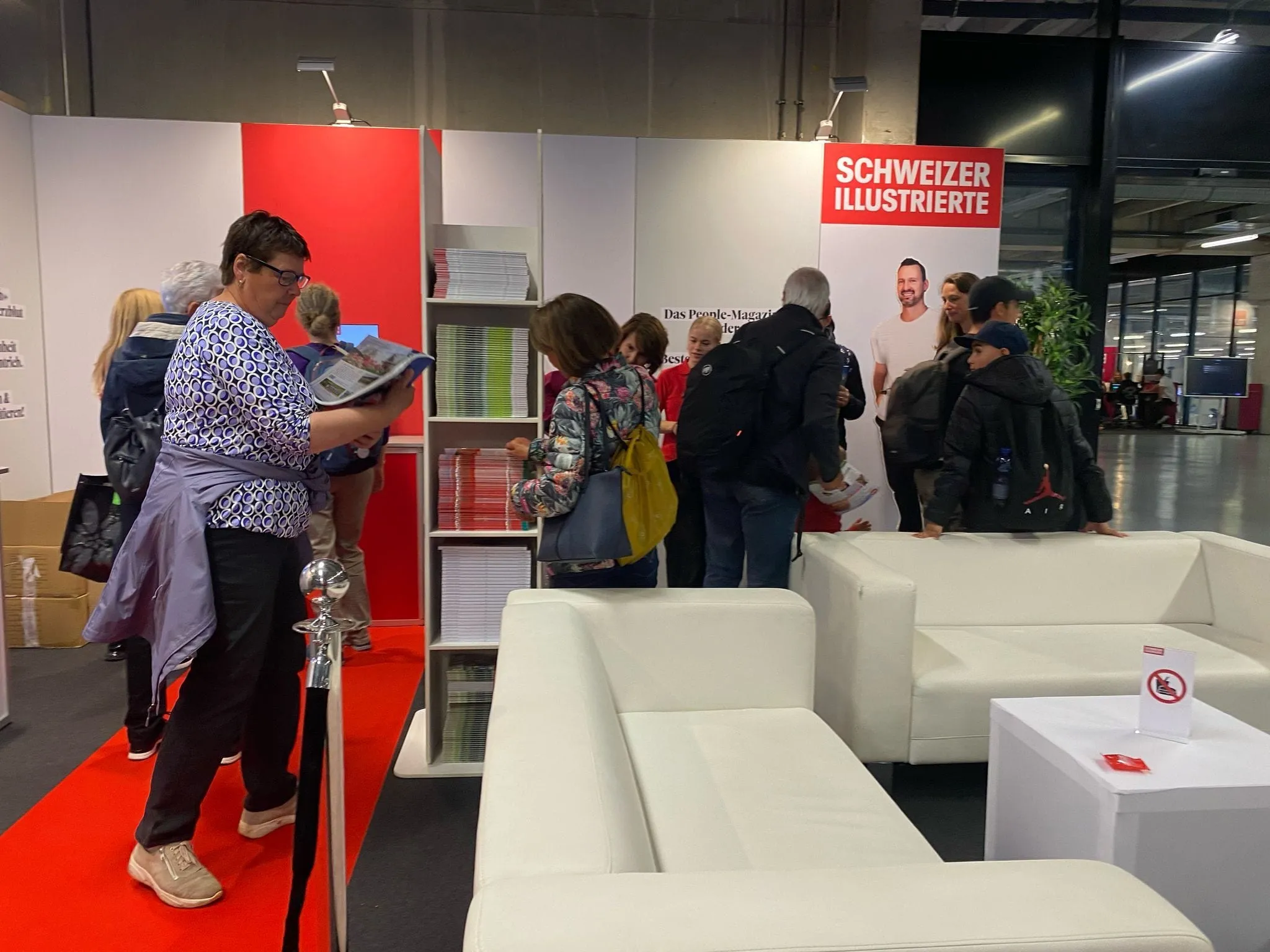 Besucherin blättert in einem Magazin am Zeitungsständer, weitere Gäste am Stand und weisse Sofas