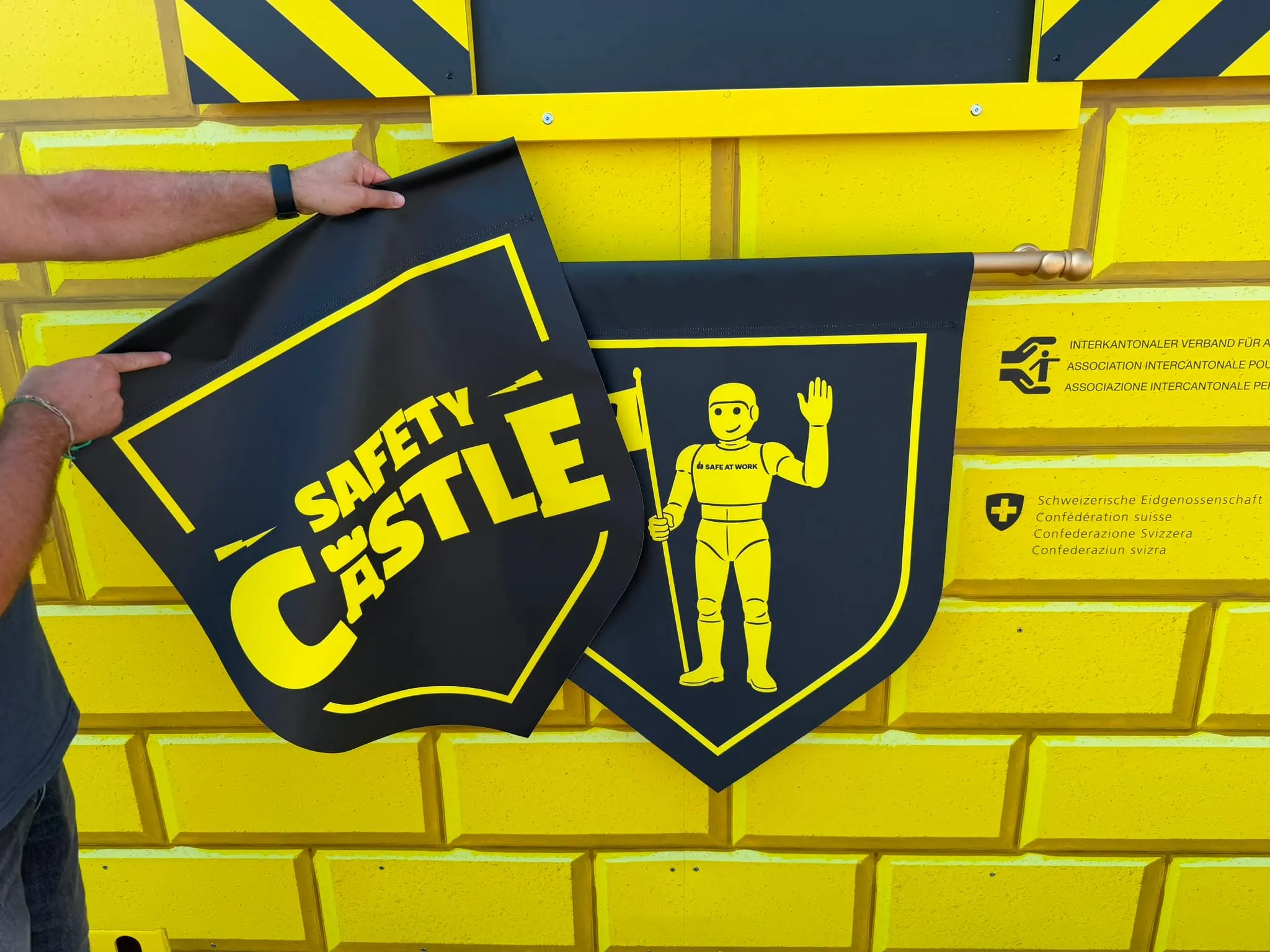 Two hands holding a black «Safety Castle» banner beside a crest with the yellow Safe at Work knight on a yellow brick-effect facade