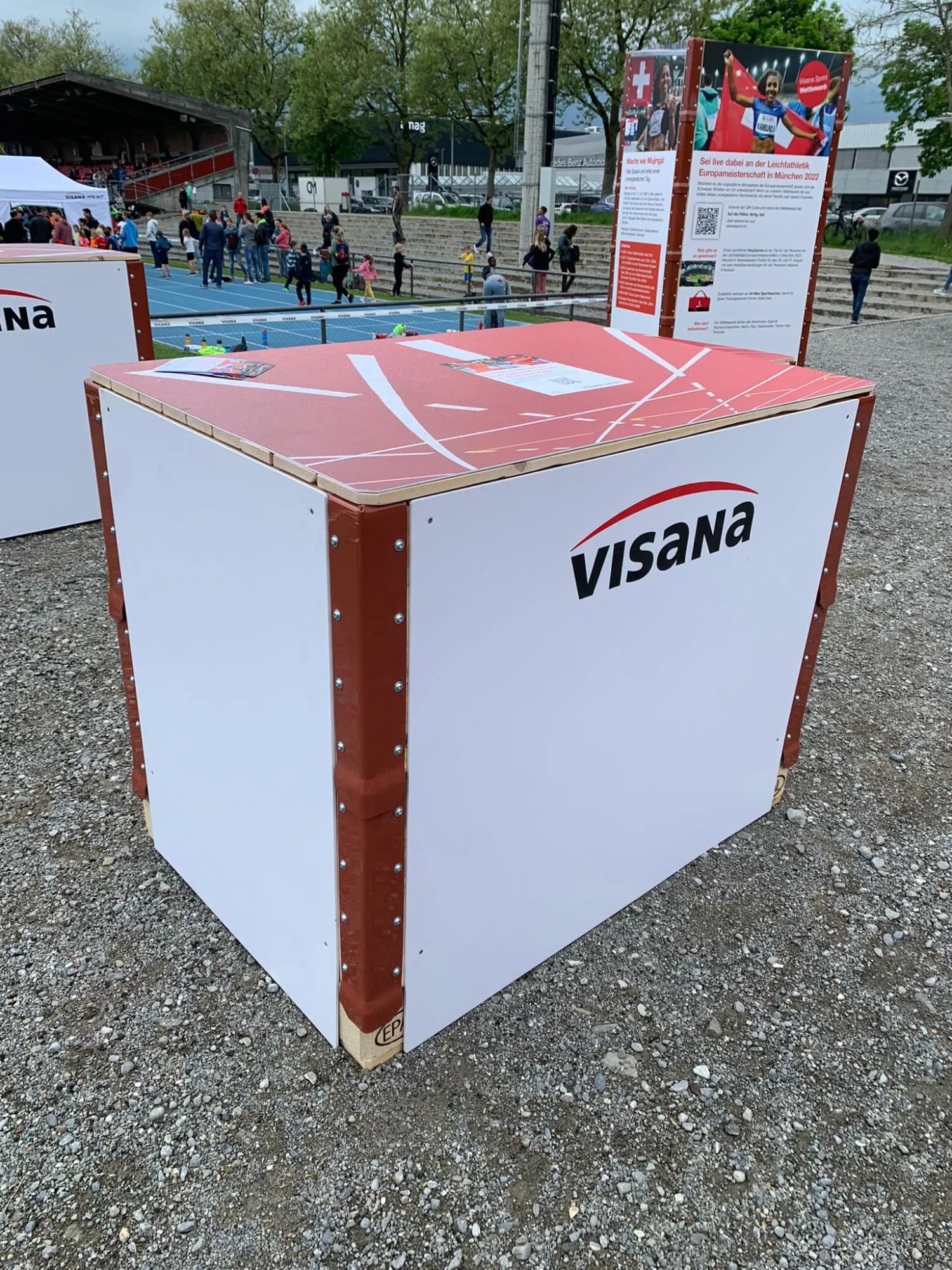 Visana-Tischbox aus Holz mit rotem Laufbahn-Deckel und weissen Seitenwänden, dahinter Infostelen und Sprintbahn