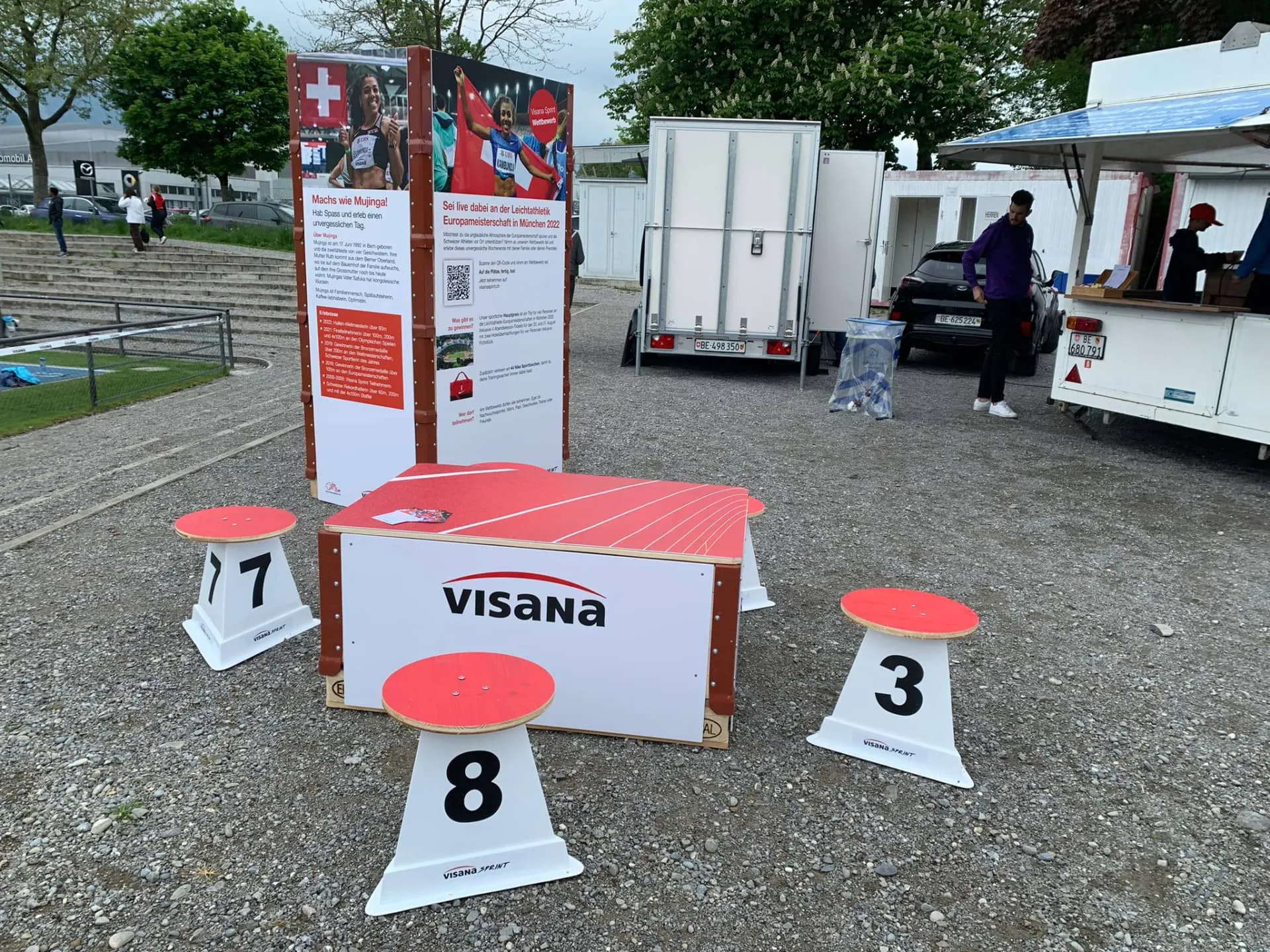 Rot-weisse Visana-Tischbox mit Laufbahn-Print, Hocker mit Startnummern 3, 7 und 8, dahinter Infostelen zu Mujinga Kambundji