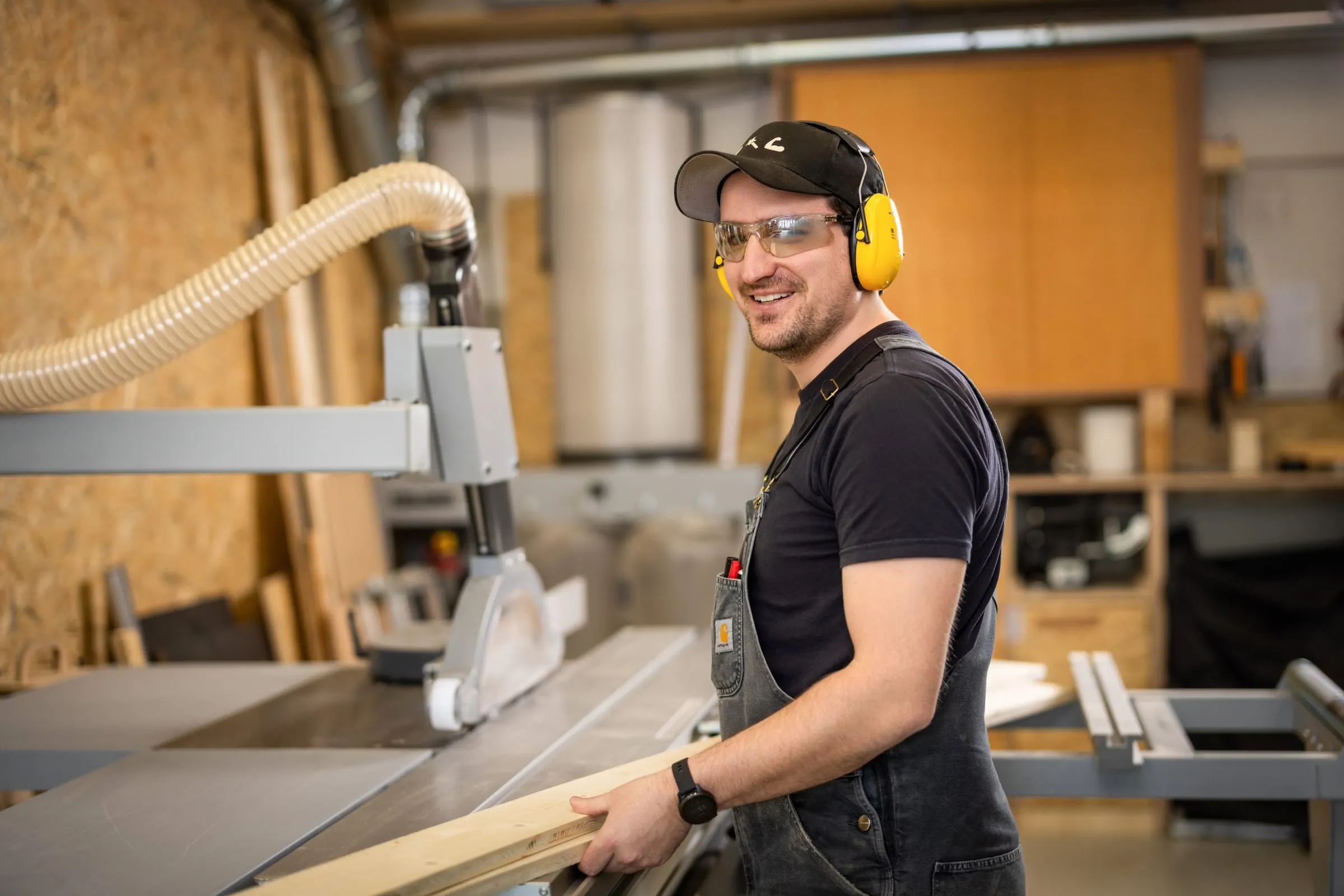Man in a cap, safety glasses and yellow ear defenders holding a wooden batten at the panel saw