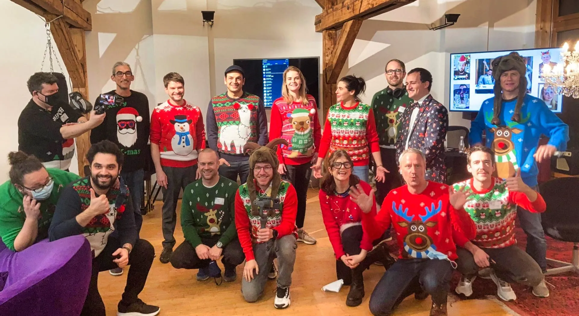 Group in ugly Christmas sweaters posing in a studio, with a video-call screen and a camera operator behind them