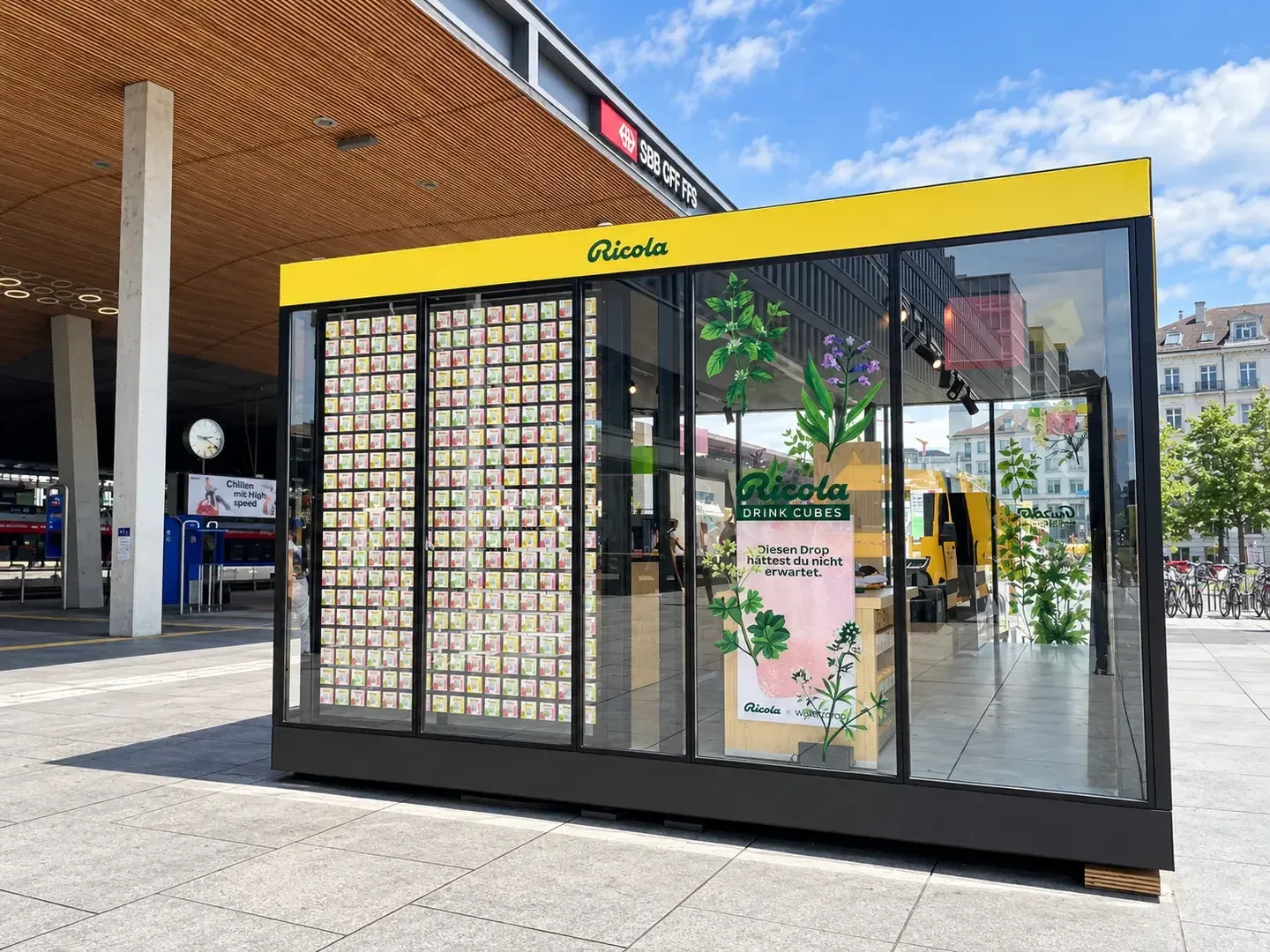 Frontansicht des Ricola-Pop-up-Shops mit Glasfassade, gelber Ricola-Blende und einer Wand aus Produktpackungen neben dem Bahnhof Zürich