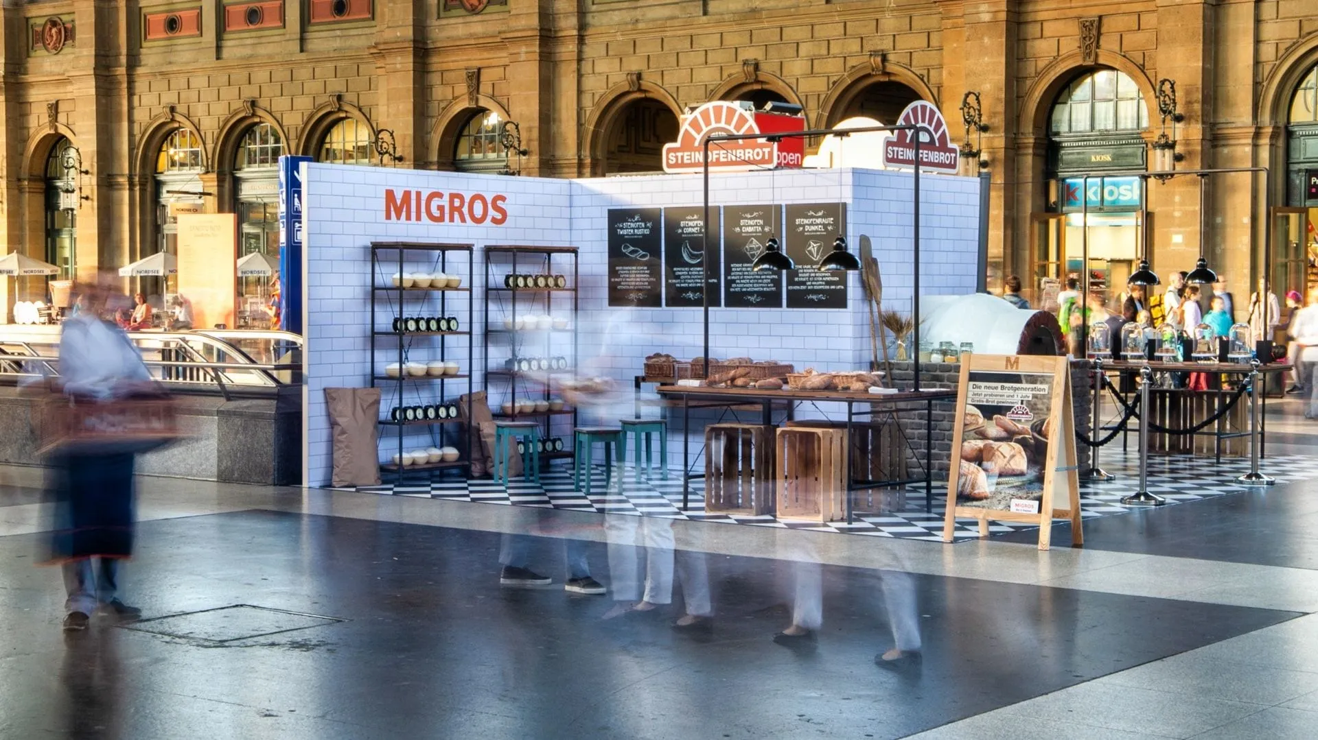 Migros stone-oven bread pop-up with a white tiled wall, shelves and a bread table in the concourse of Zürich main station