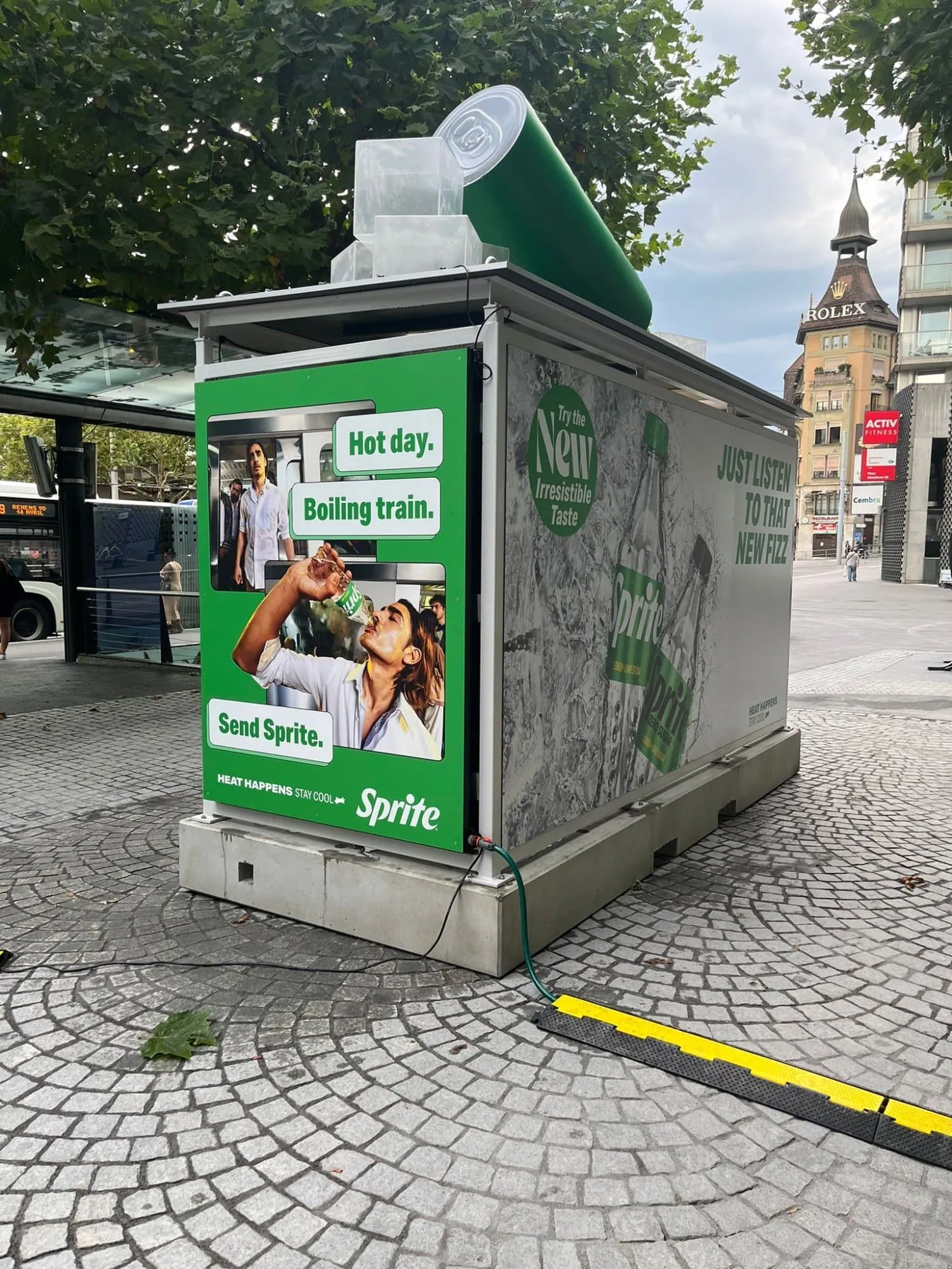 Rückseite der Sprite-Haltestelle mit Sprite-Zero-Flaschenmotiv und liegender XXL-Dose vor einer Busstation in Lausanne