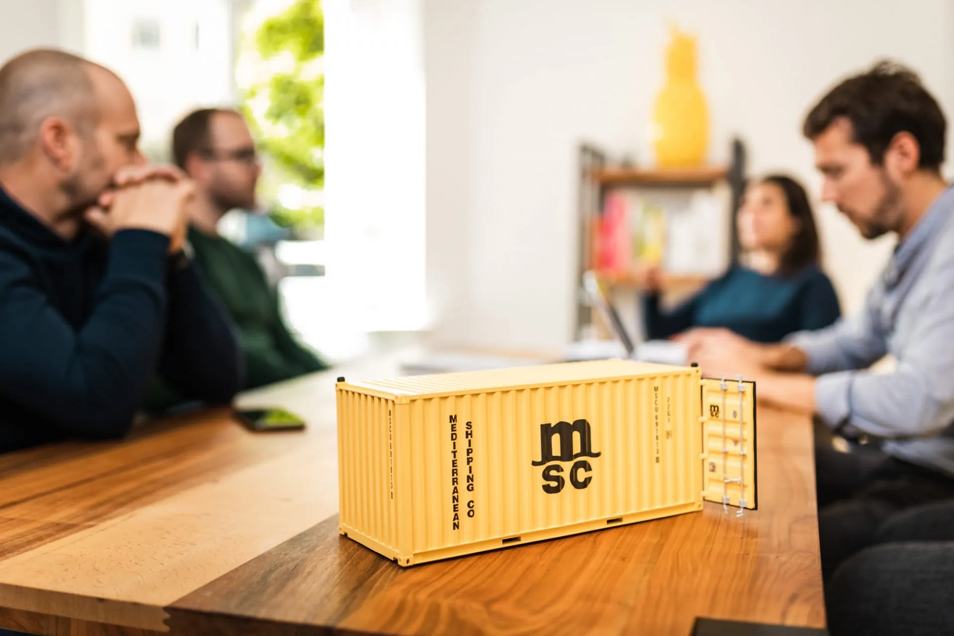 Gelbes MSC-Container-Modell auf einem Holztisch, dahinter vier Personen in einer Besprechung