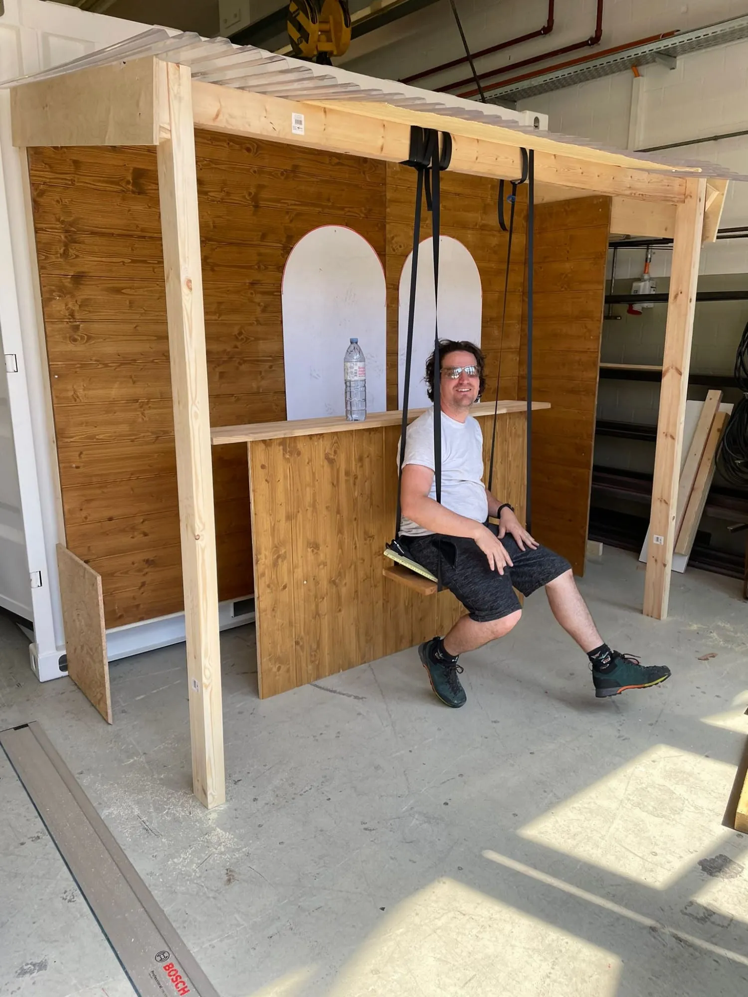 Mann testet den Schaukelsitz an der Holz-Bar mit Pergola in der Werkstatt