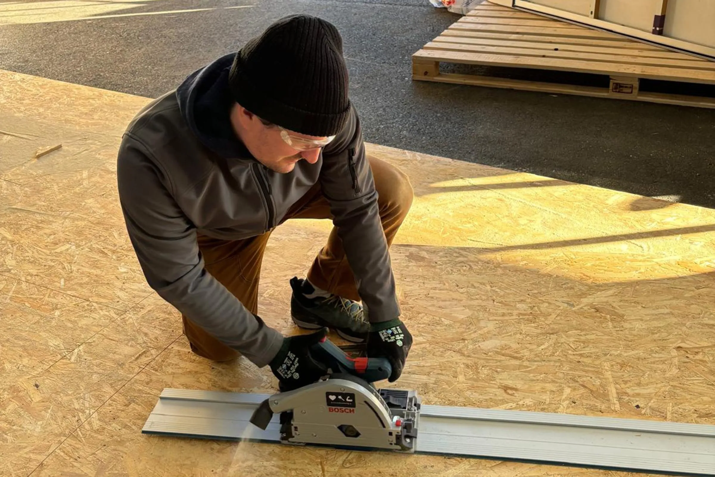 Man in a beanie and safety glasses kneeling on an OSB board, cutting with a plunge saw on a guide rail