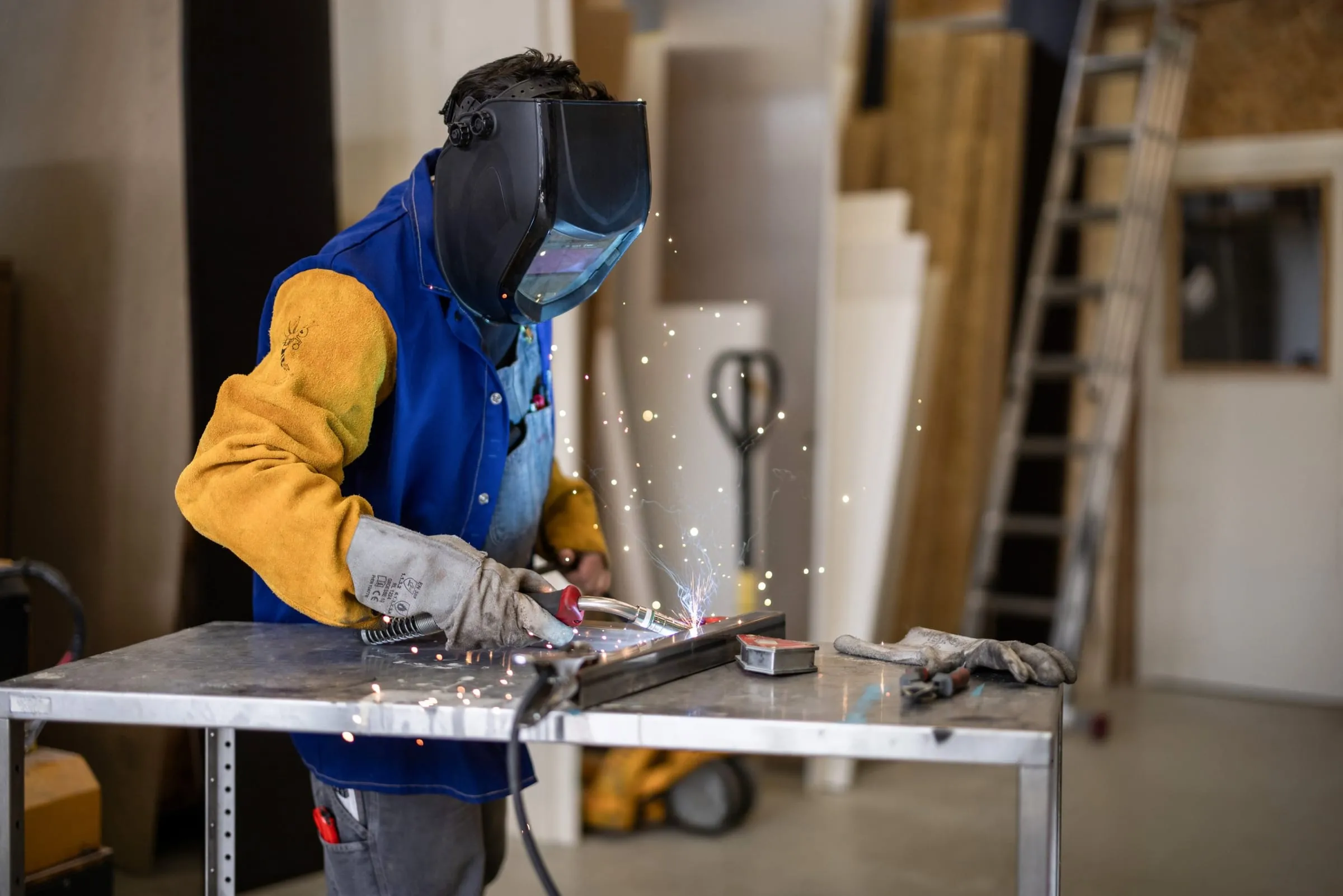 Person in a welding helmet and a blue and yellow protective jacket welding a metal profile, sparks flying