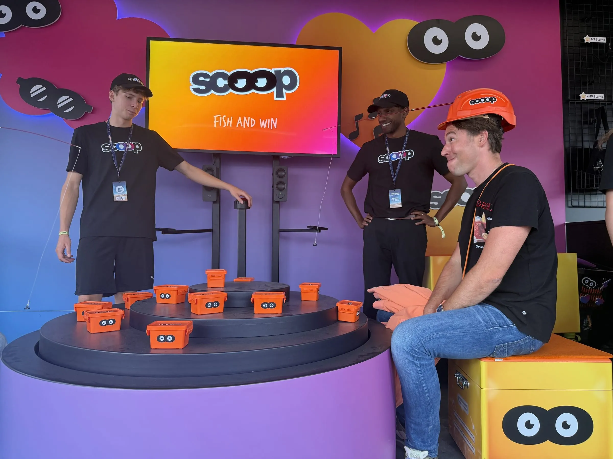 Man in an orange scoop helmet sitting at the rotating platform with orange loot boxes, two promoters at the buzzer in front of the «Fish and Win» screen