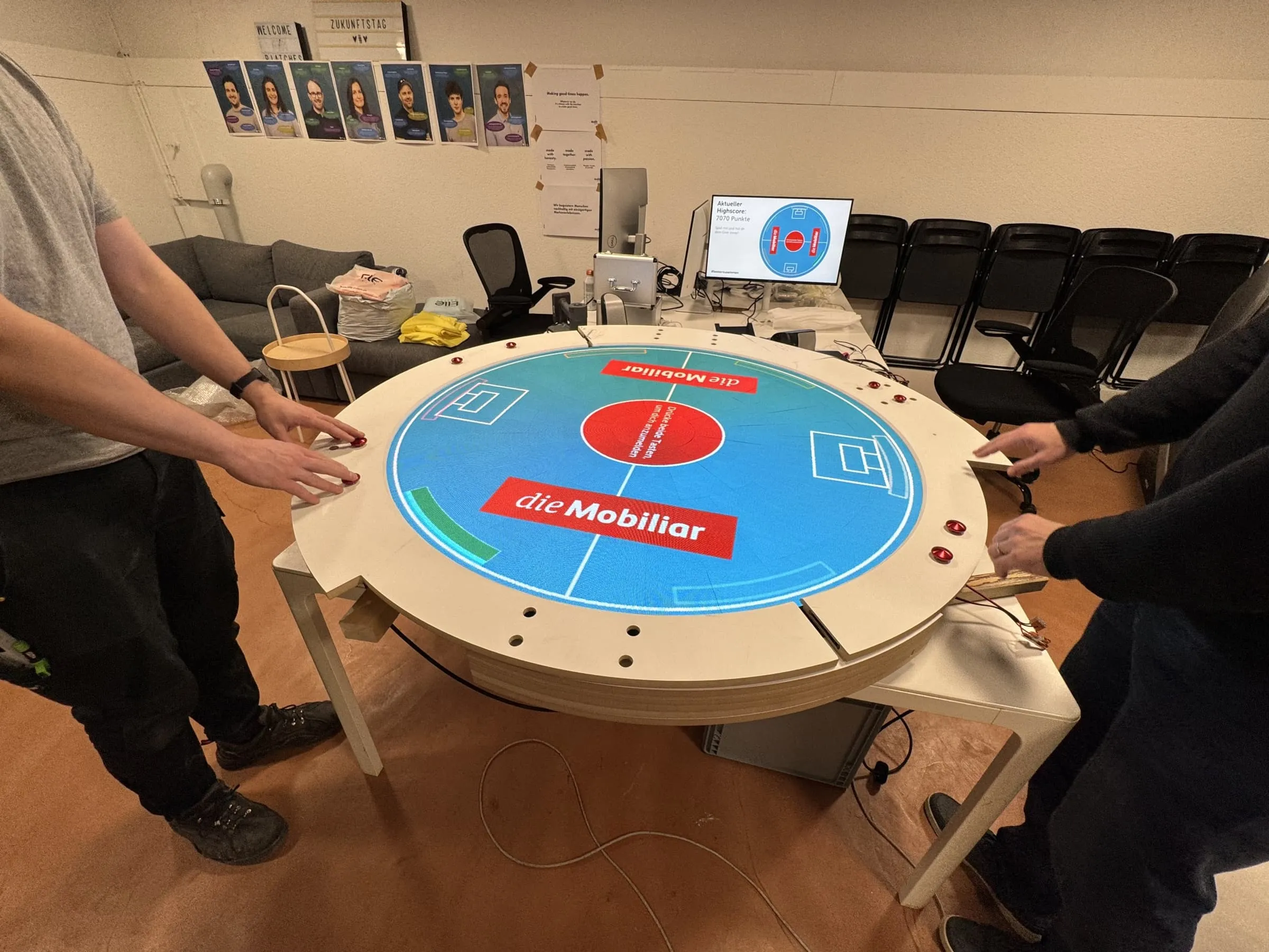 Prototyp des runden Spieltischs im Büro, der LED-Screen zeigt das blaue Pong-Spielfeld mit Mobiliar-Logos, zwei Personen an den roten Arcade-Buttons