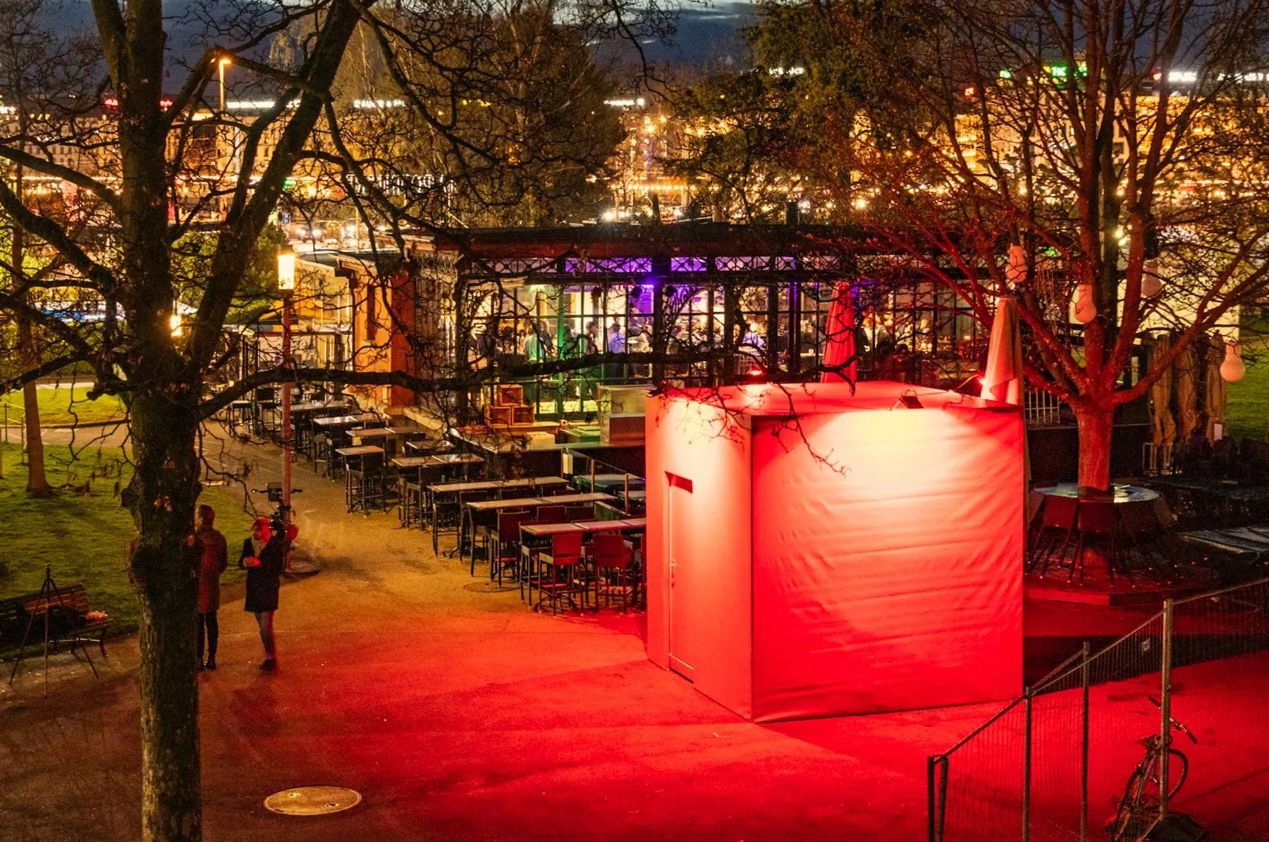 Screenshot of a red-lit cube at night beside a bar terrace, with city lights in the background