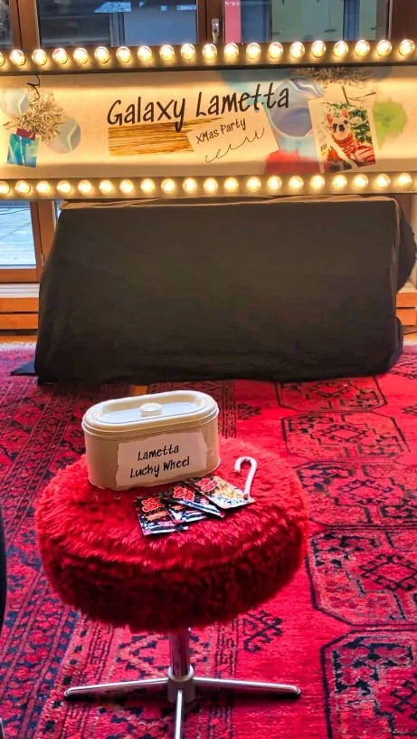 Roter Plüschhocker mit Box «Lametta Lucky Wheel» und Karten vor Leuchtschild «Galaxy Lametta XMas Party»
