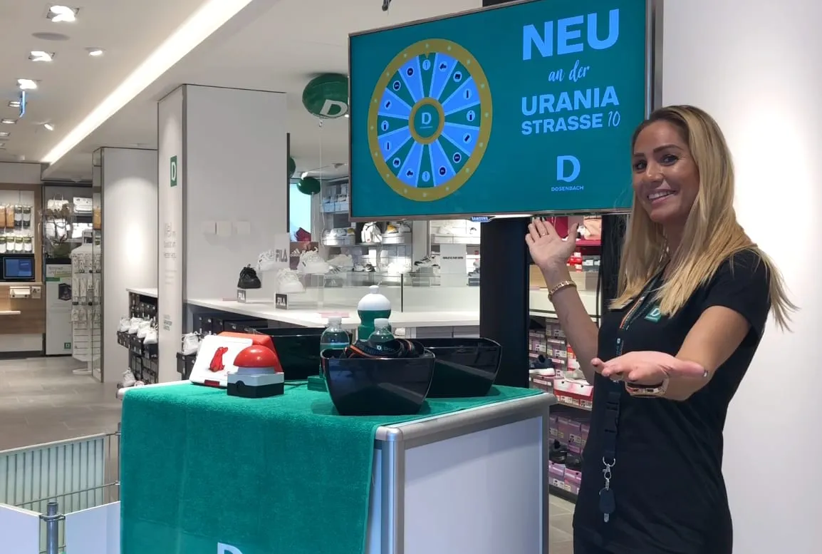 Promotorin zeigt auf den Bildschirm mit Glücksrad, davor ein roter Buzzer auf grünem Tischtuch im Schuhgeschäft