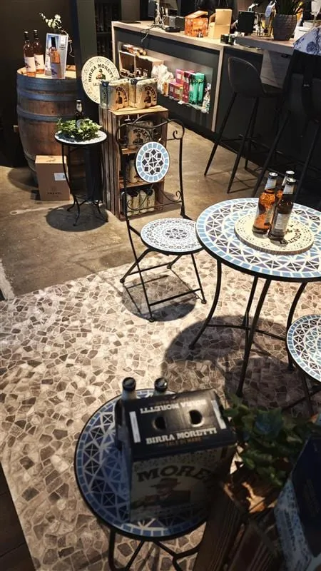 Mosaiktische und Klappstühle auf dem Steinmosaik-Boden im Laden, dazu Bierkarton und Holzfass mit Flaschen