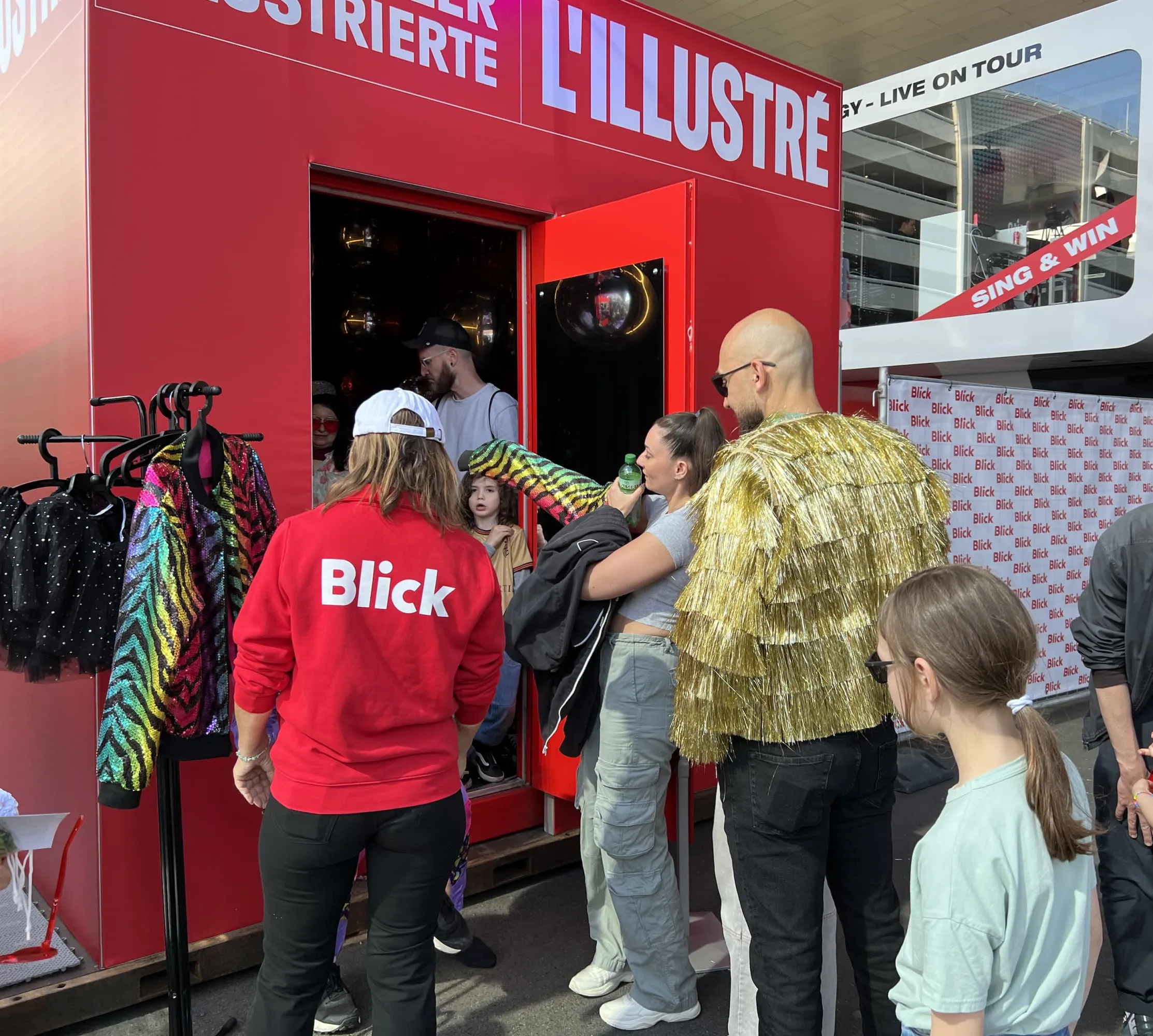 Roter SI-Cube mit Aufschrift «L'Illustré», Kleiderständer mit Paillettenjacken und Besuchende am Eingang, ein Mann im goldenen Fransenmantel