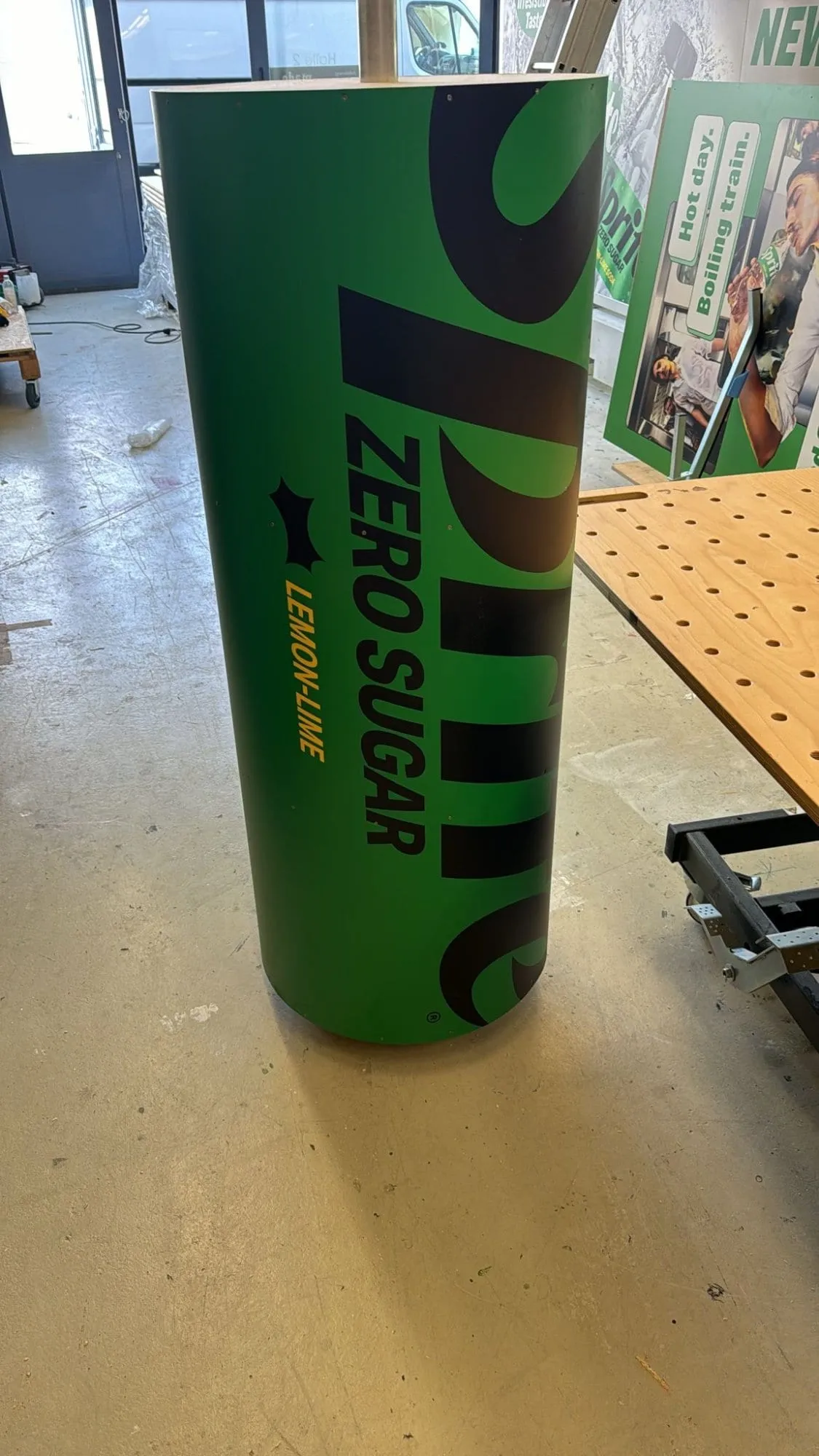 Überdimensionale grüne Sprite-Zero-Sugar-Dose steht auf dem Werkstattboden, dahinter Plakatwände