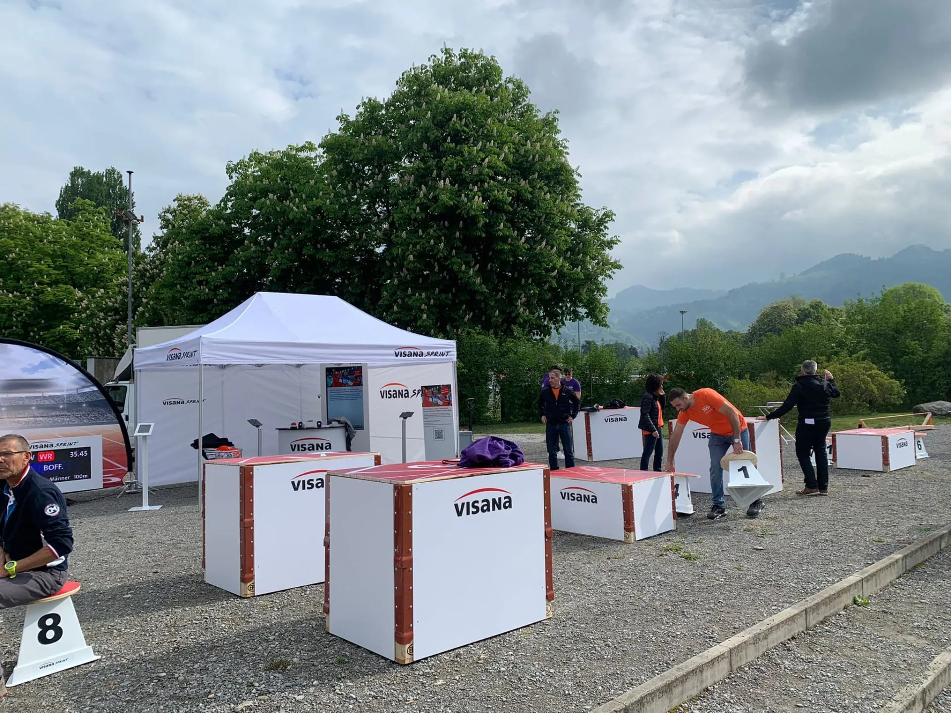 Visana-Sprint-Zelt mit Bildschirmen, davor rot-weisse Visana-Möbel aus Holzkisten und nummerierte Hocker auf Kies
