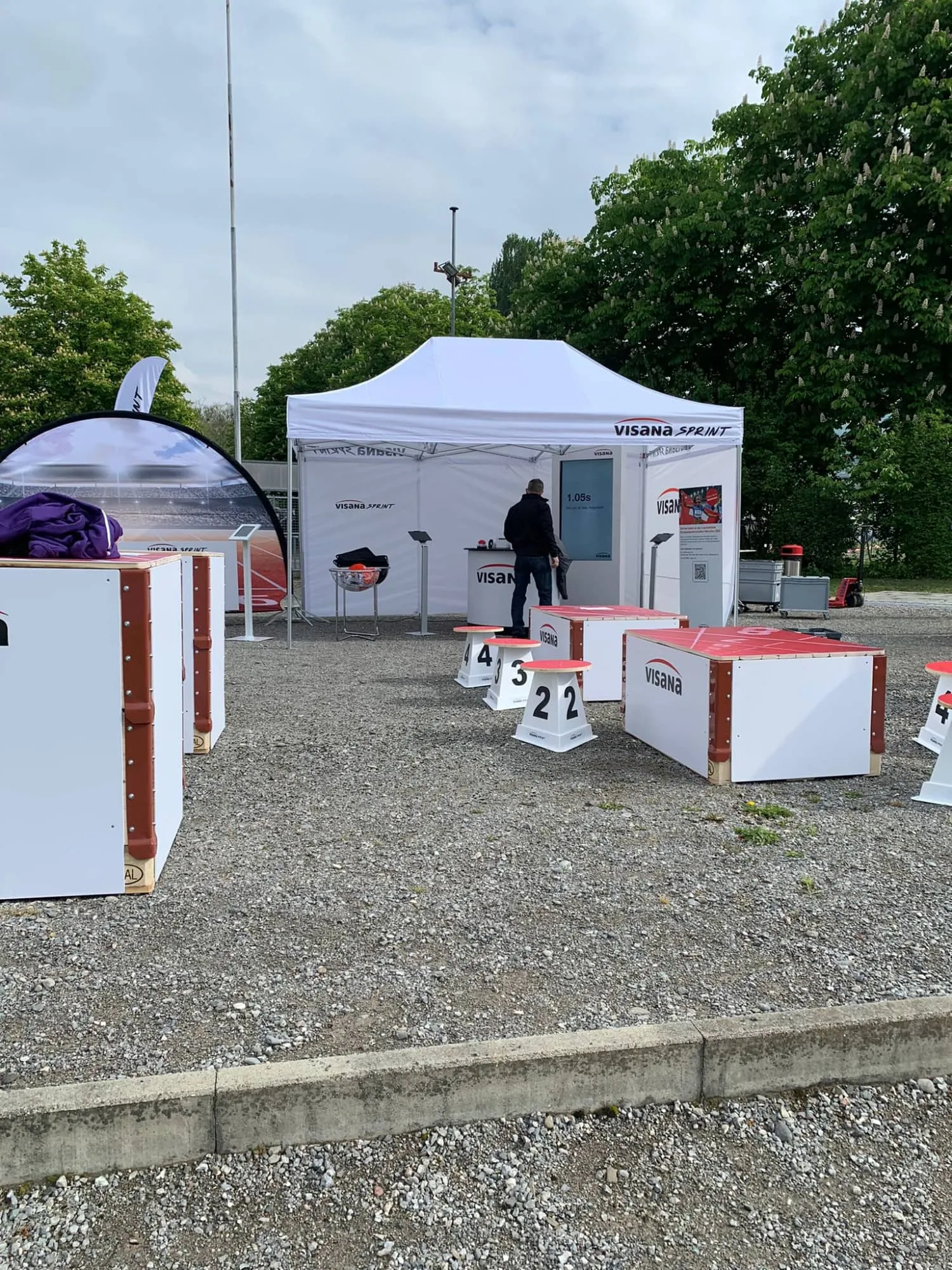 Visana-Piazza mit Zelt, Bildschirm, nummerierten Hockern und Visana-Boxen auf einem Kiesplatz