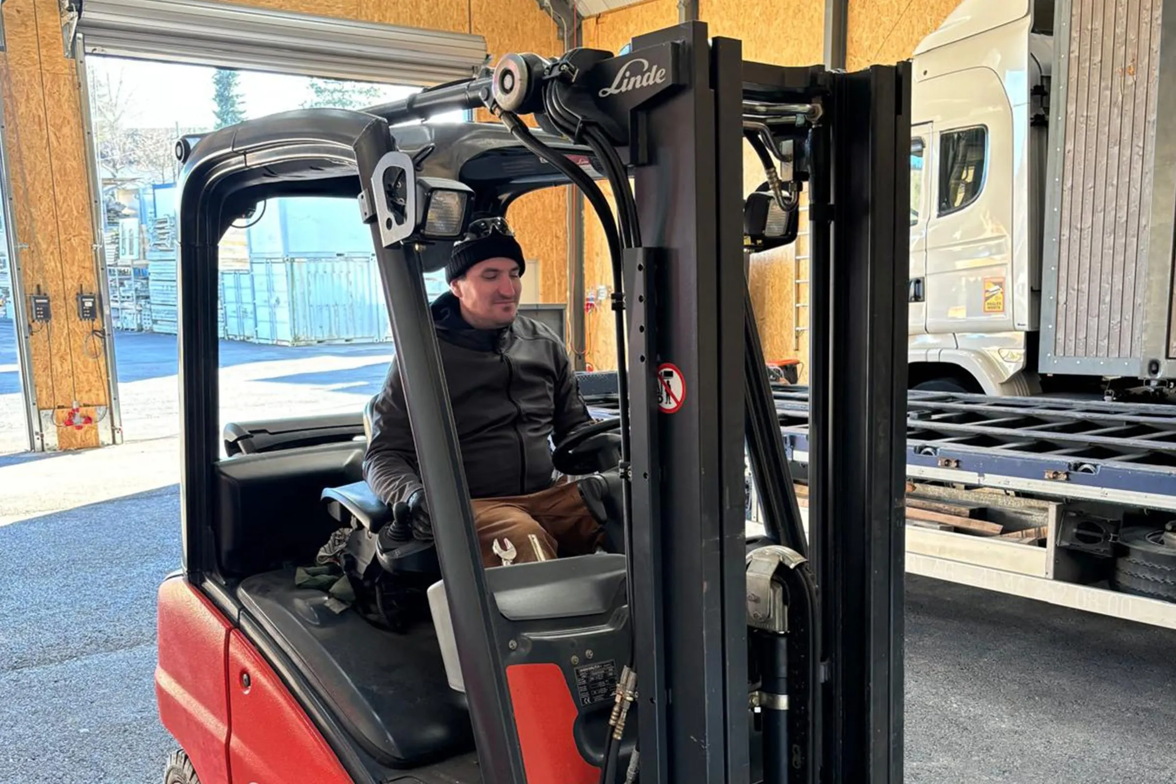 Man in a beanie at the wheel of a red Linde forklift inside the hall, a lorry behind him