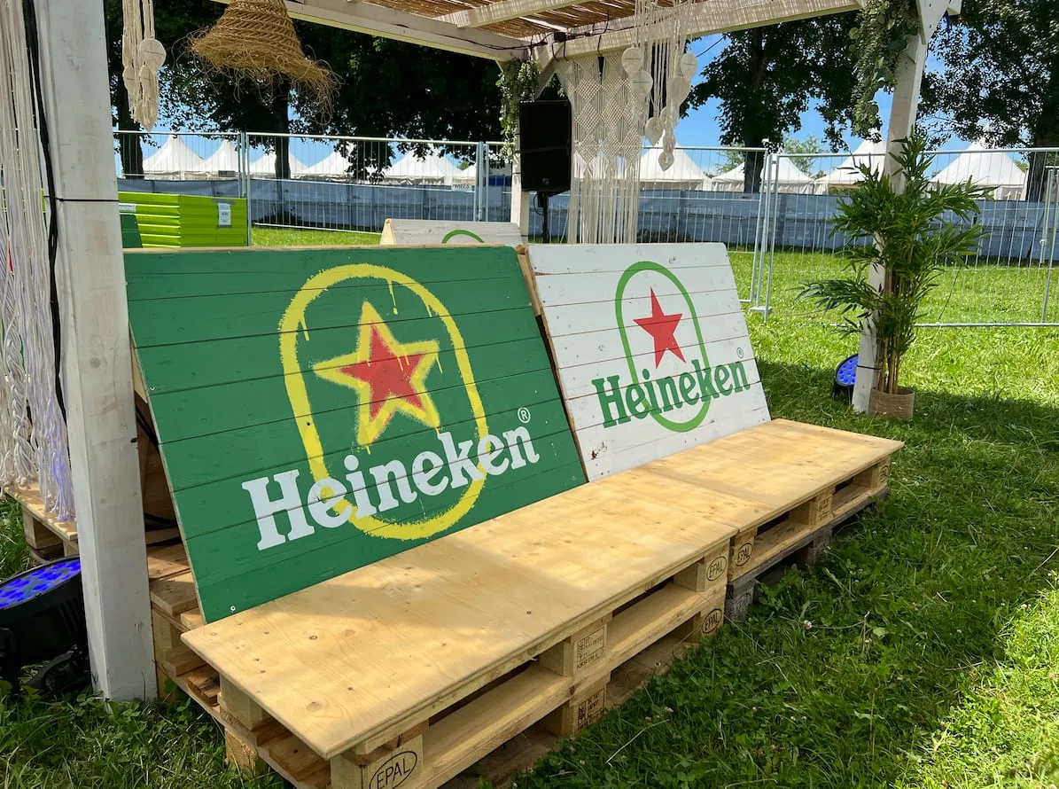 Pallet bench under a pergola with two wooden Heineken signs, a green logo and red star, with festival tents behind