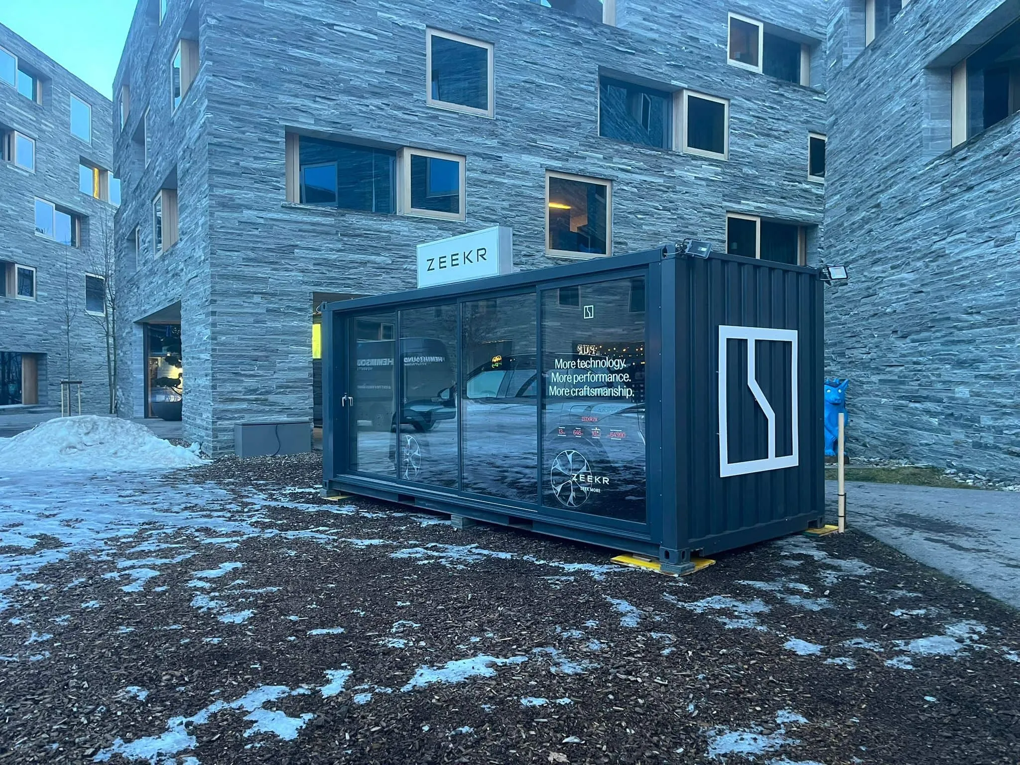 Dark Zeekr container with a glass front, white illuminated sign and end-wall logo in front of stone facades in Laax, with patches of snow