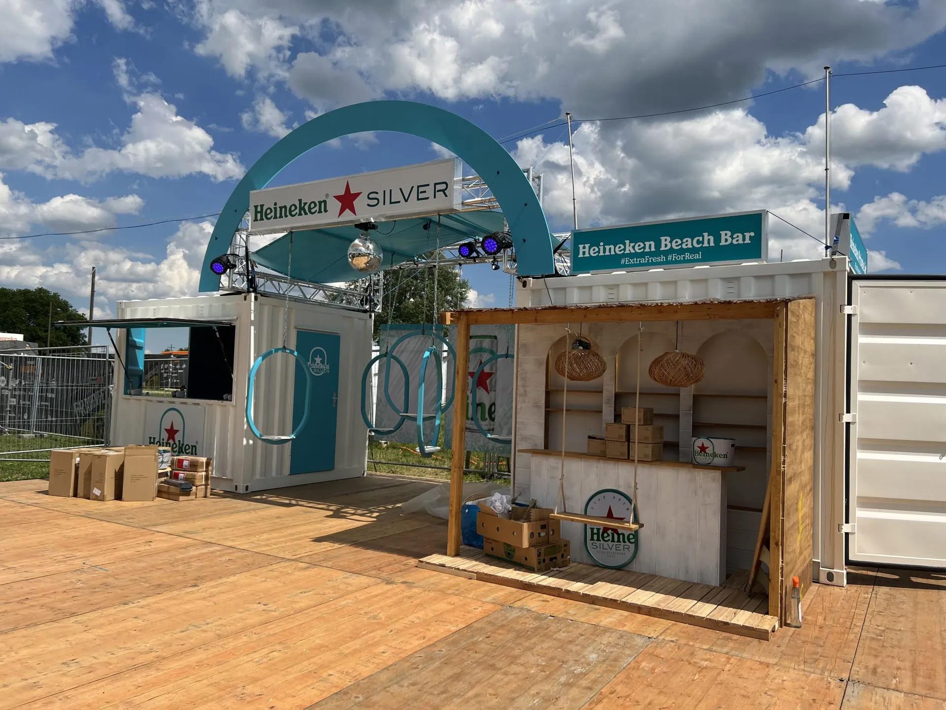 Türkiser Heineken-Silver-Bogen mit Discokugel über den Containern, davor Ringschaukeln und die Beach Bar