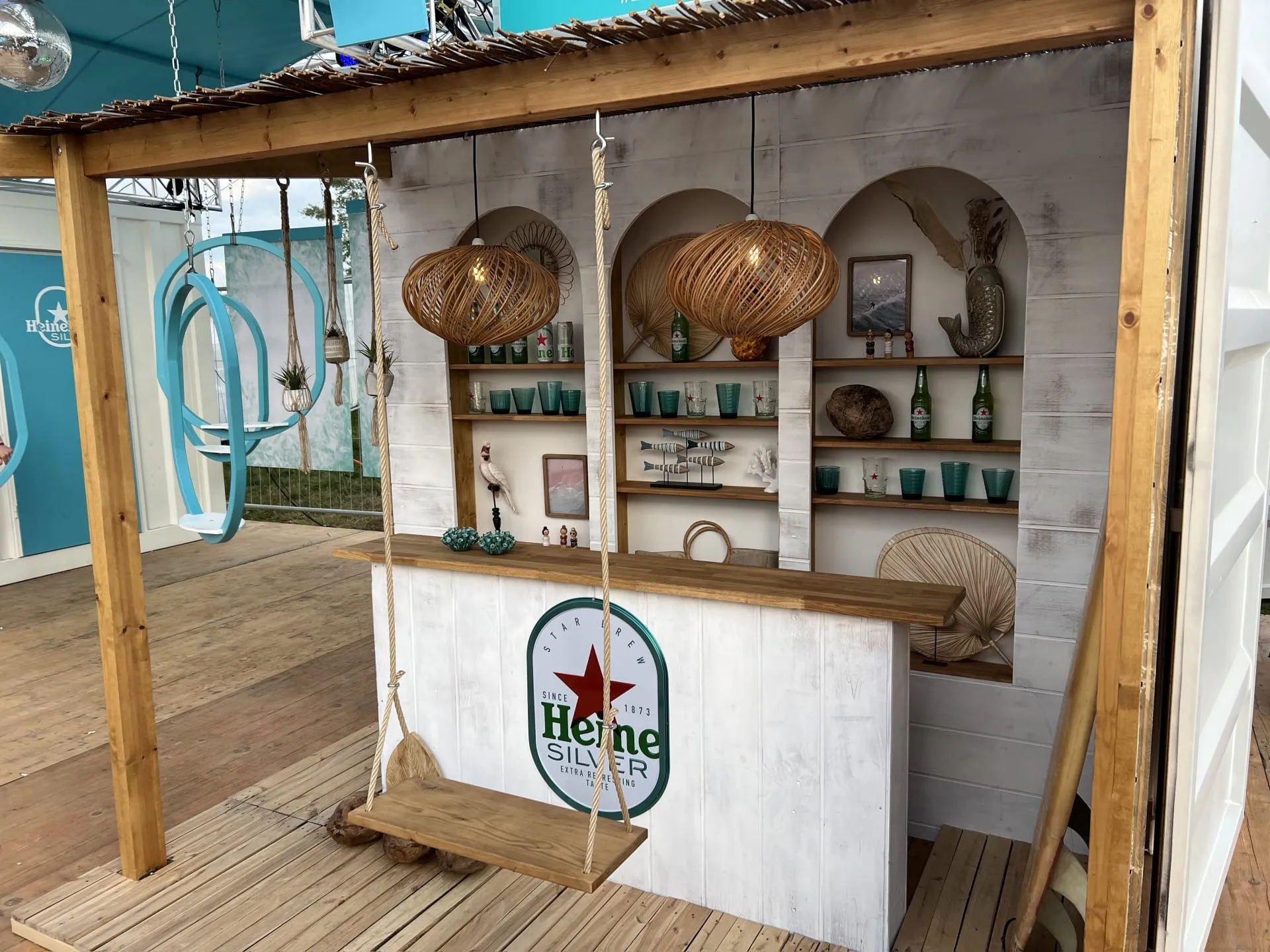 White-painted wooden Heineken Beach Bar with a swing seat, rattan lamps and shelves, beside turquoise ring swings