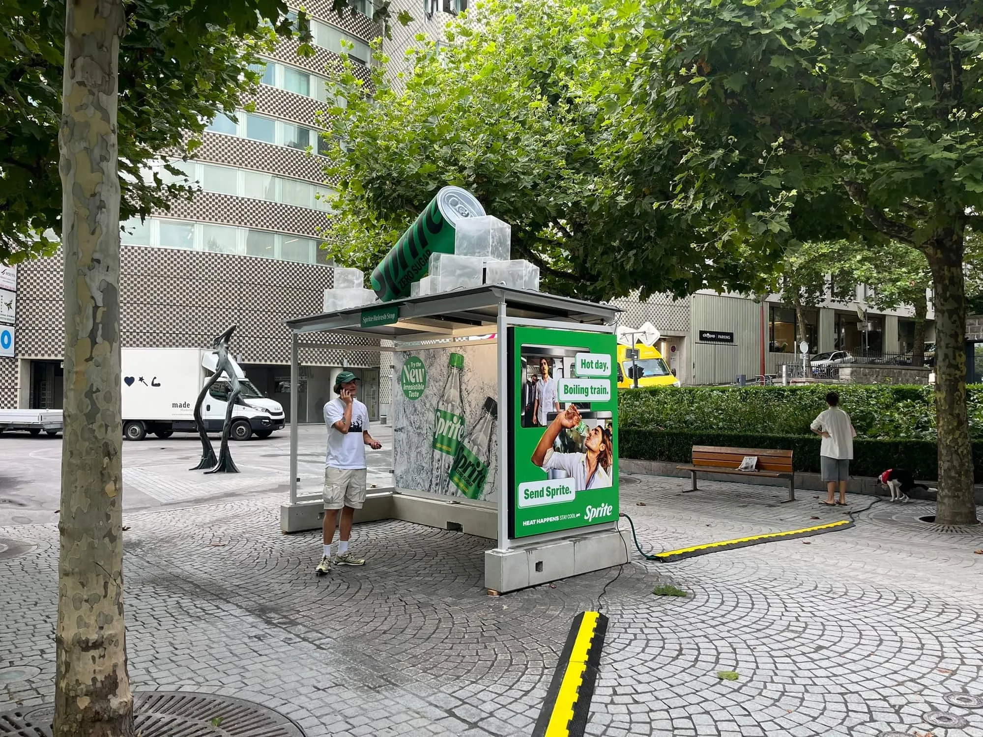 Sprite bus stop with an XXL can and ice cubes on the roof, on a paved square under plane trees, with a man on the phone beneath it