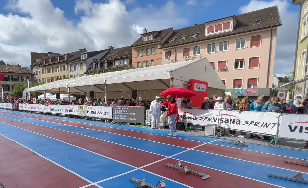 Blue sprint track with starting blocks, Visana Sprint barriers and spectators in front of a marquee and buildings in Winterthur