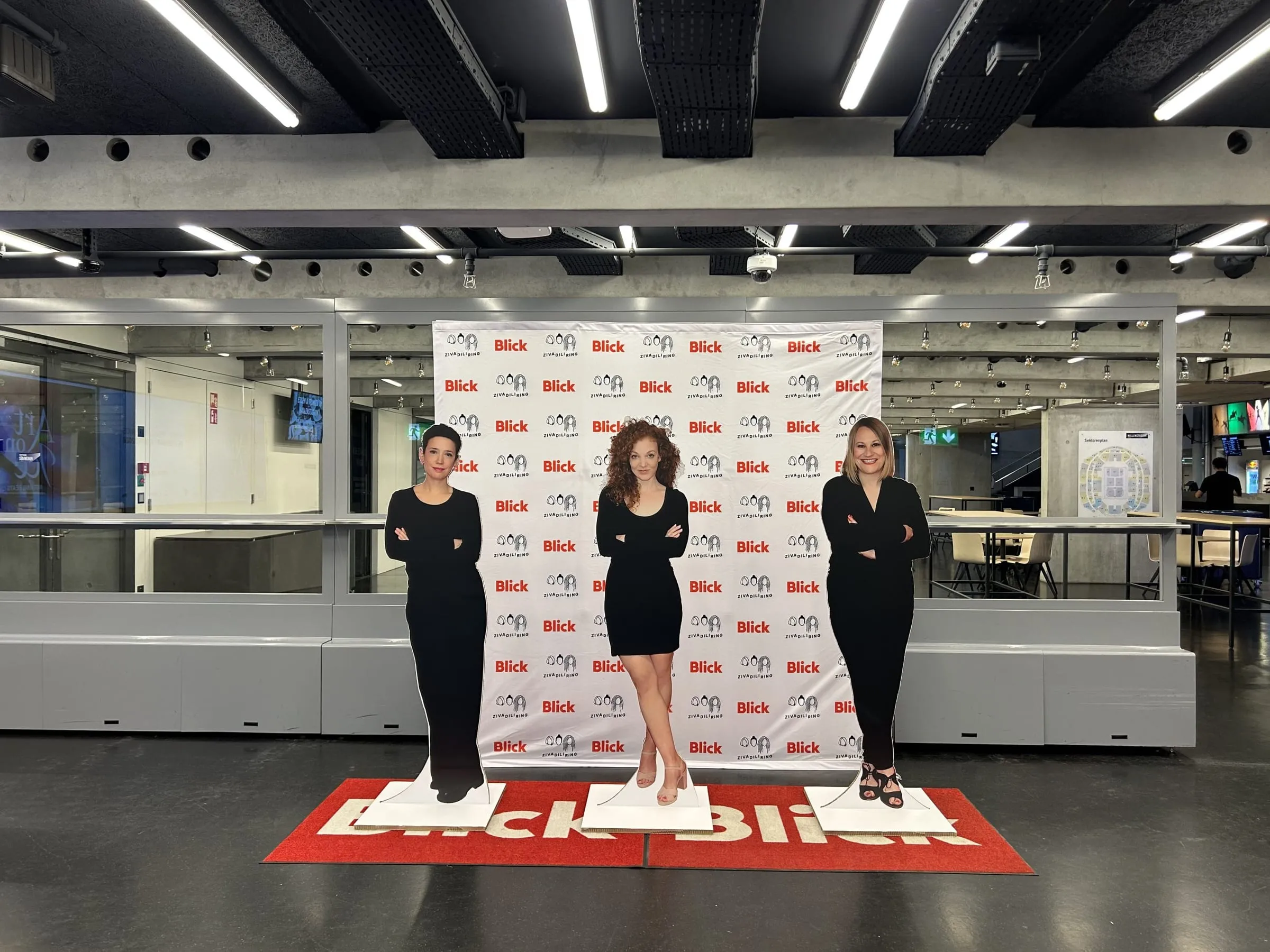 Three life-size cardboard cutouts of the Zivadiliring podcast hosts in front of a Blick logo wall on a red Blick carpet
