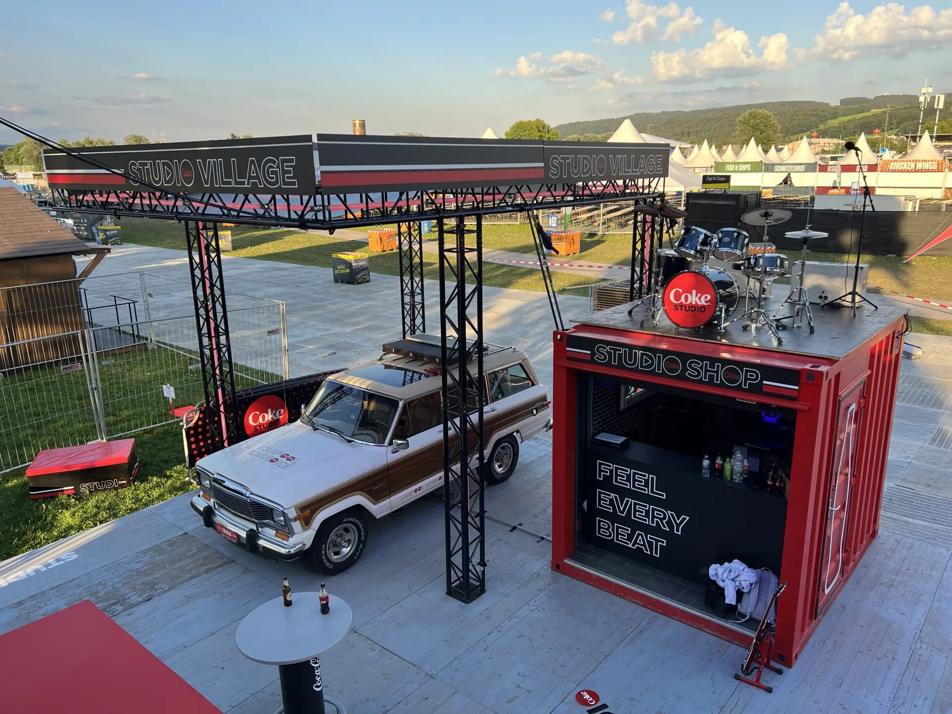 View from above of the Coke Studio Village: Carpool Karaoke Jeep beneath a truss roof, red container with Studio Shop and a drum kit