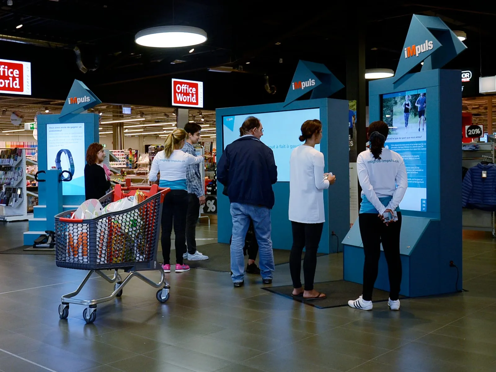 Blue iMpuls stations with screens in a shopping centre, with visitors and promotional staff in front